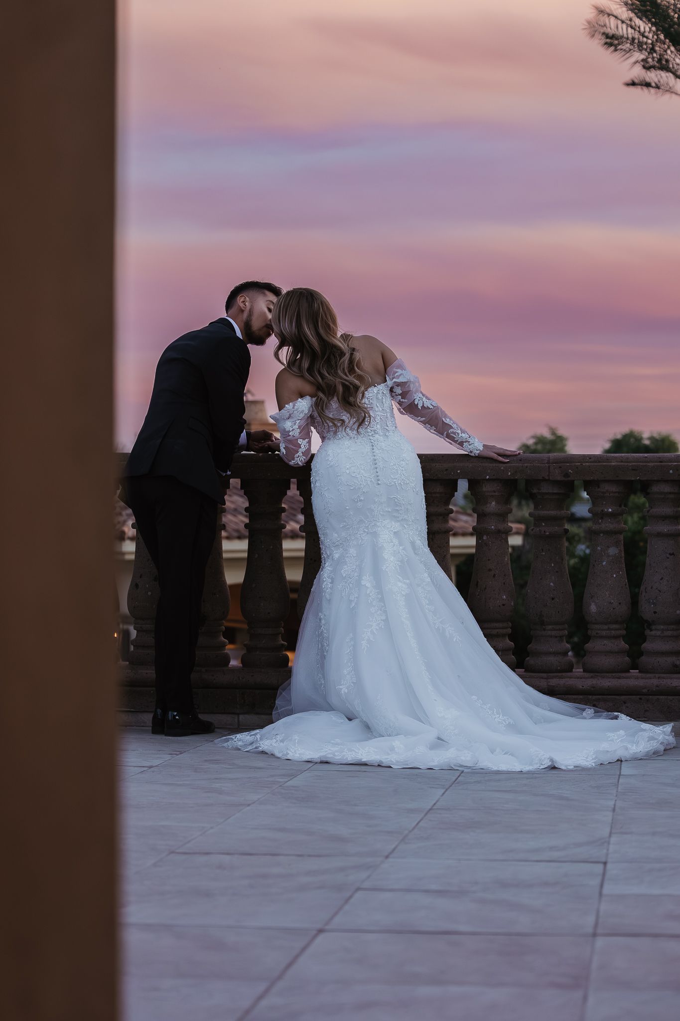 Bride and groom sharing a kiss on balcony at sunset with stunning pink and purple sky.