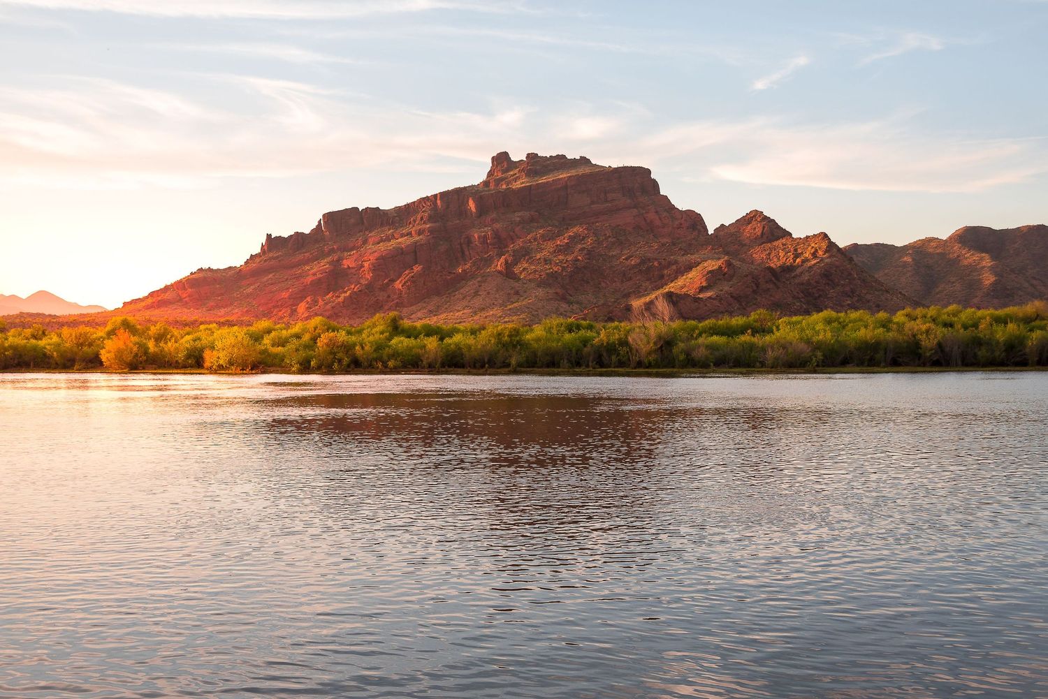 Serene Colorado River reflecting red rock mountains and lush green trees at golden sunset in Arizona desert landscape.