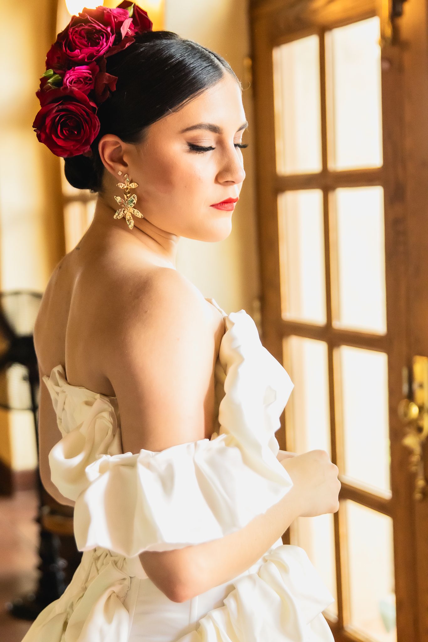 Beautiful bride with red roses in her hair wearing a white off-shoulder gown and gold earrings by a sunlit window.