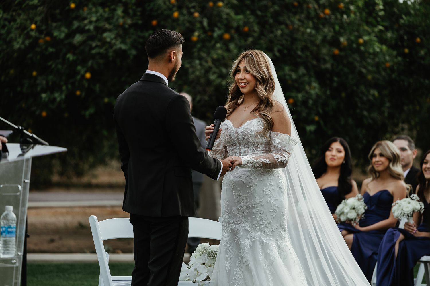 Bride in lace gown smiles at groom during outdoor wedding ceremony with guests seated nearby.