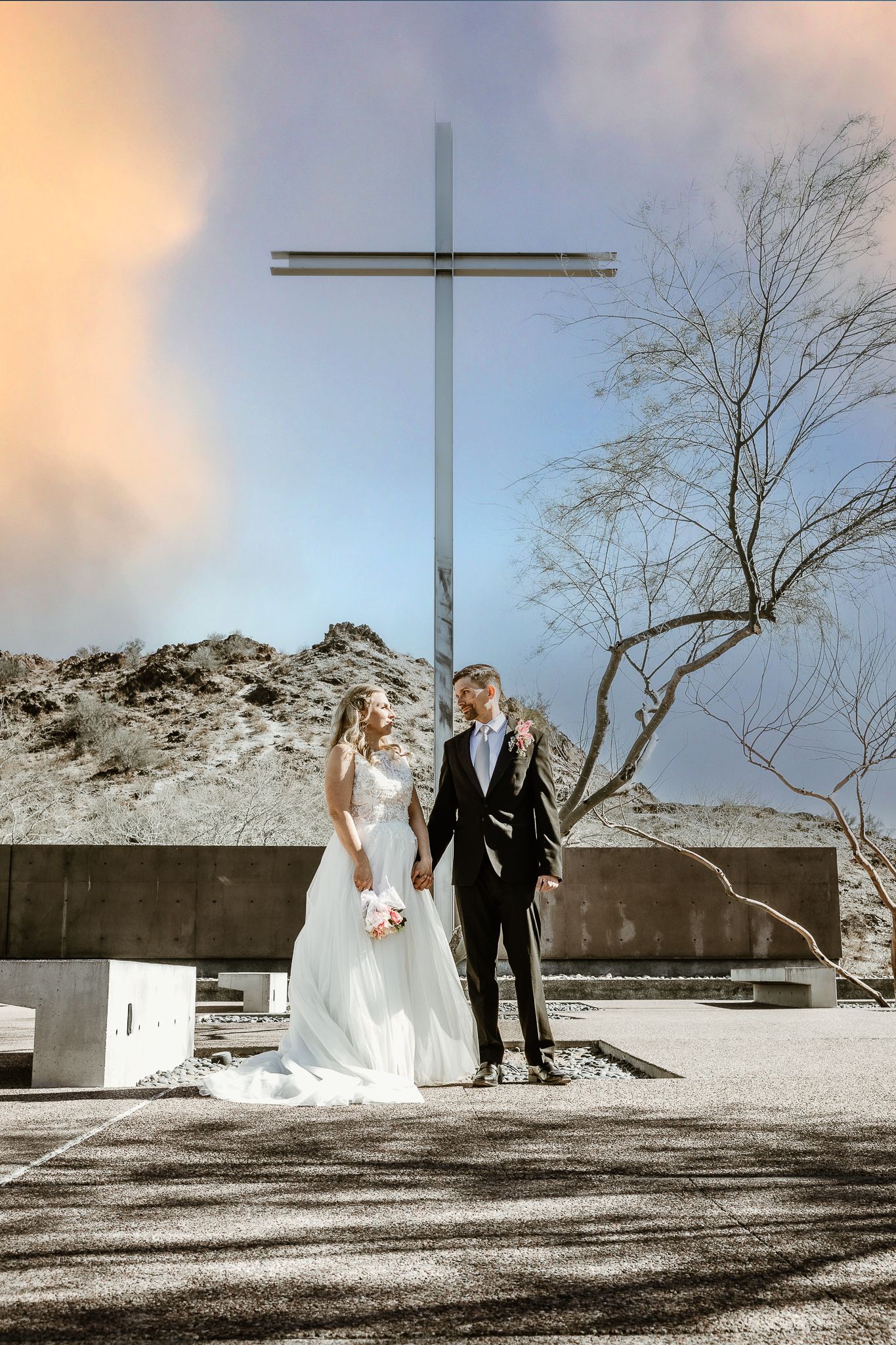 Bride and groom hold hands before large steel cross at desert wedding venue under dramatic sky.