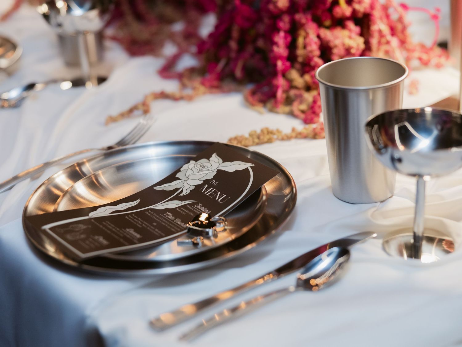 Elegant wedding table setting with dark metallic charger plate, menu card, silver goblet, and pink floral centerpiece.