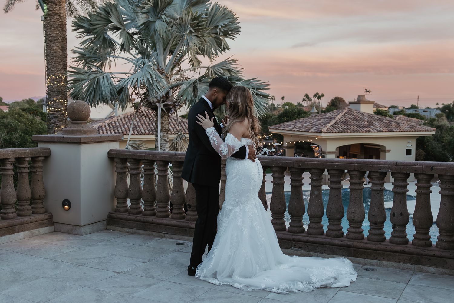 Bride and groom embracing on terrace at sunset with palm trees and rooftop views behind them.