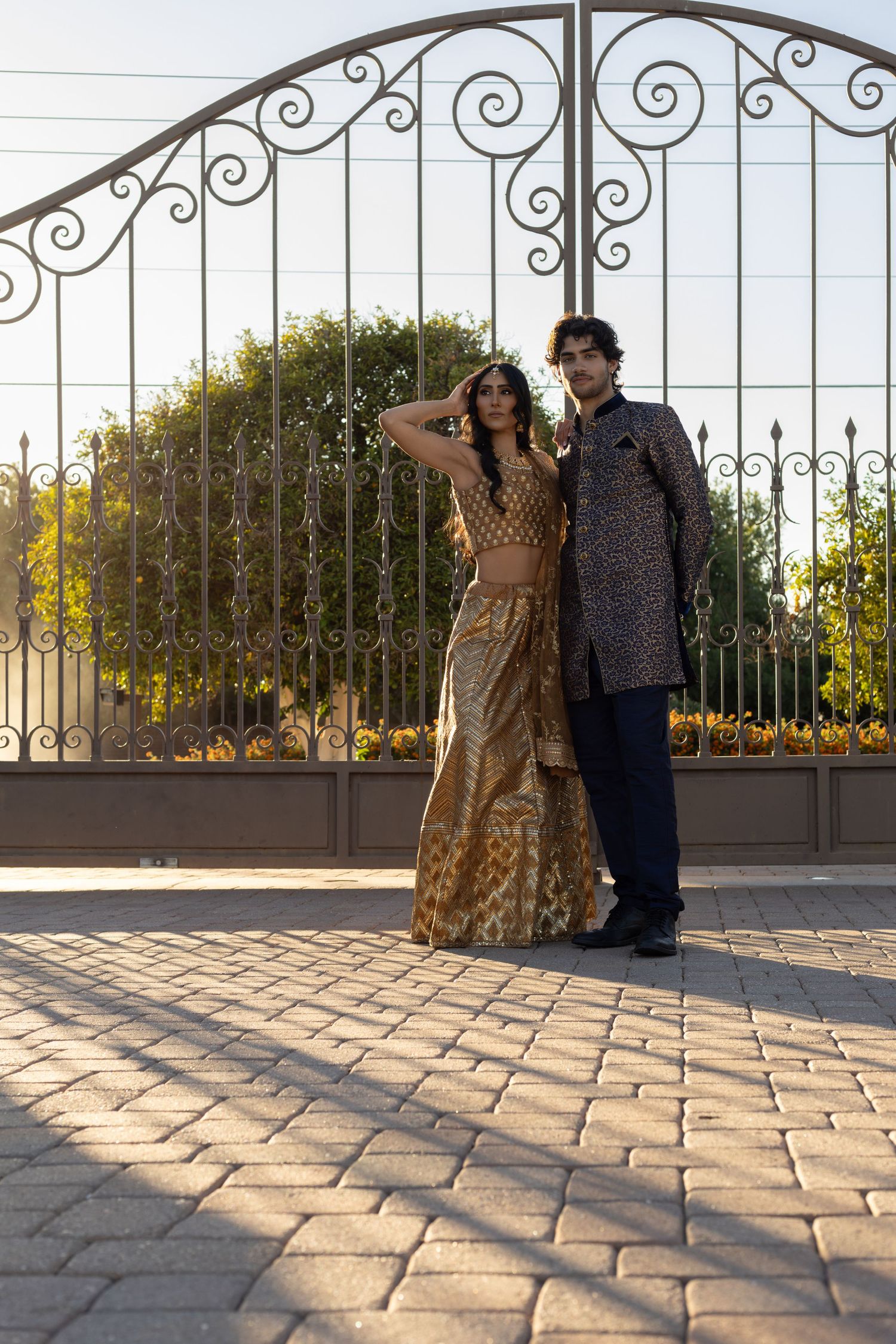 Couple in traditional Indian wedding attire posing before ornate iron gates at golden hour.