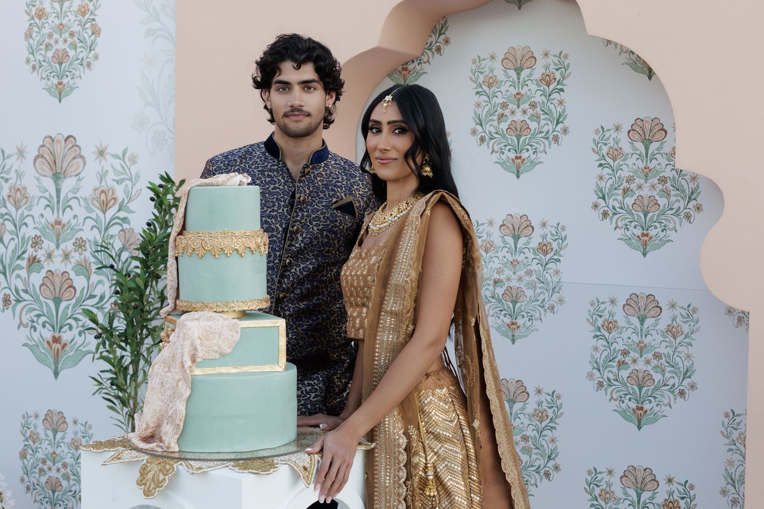 Couple in Indian wedding attire stands beside a three-tier mint and gold cake.