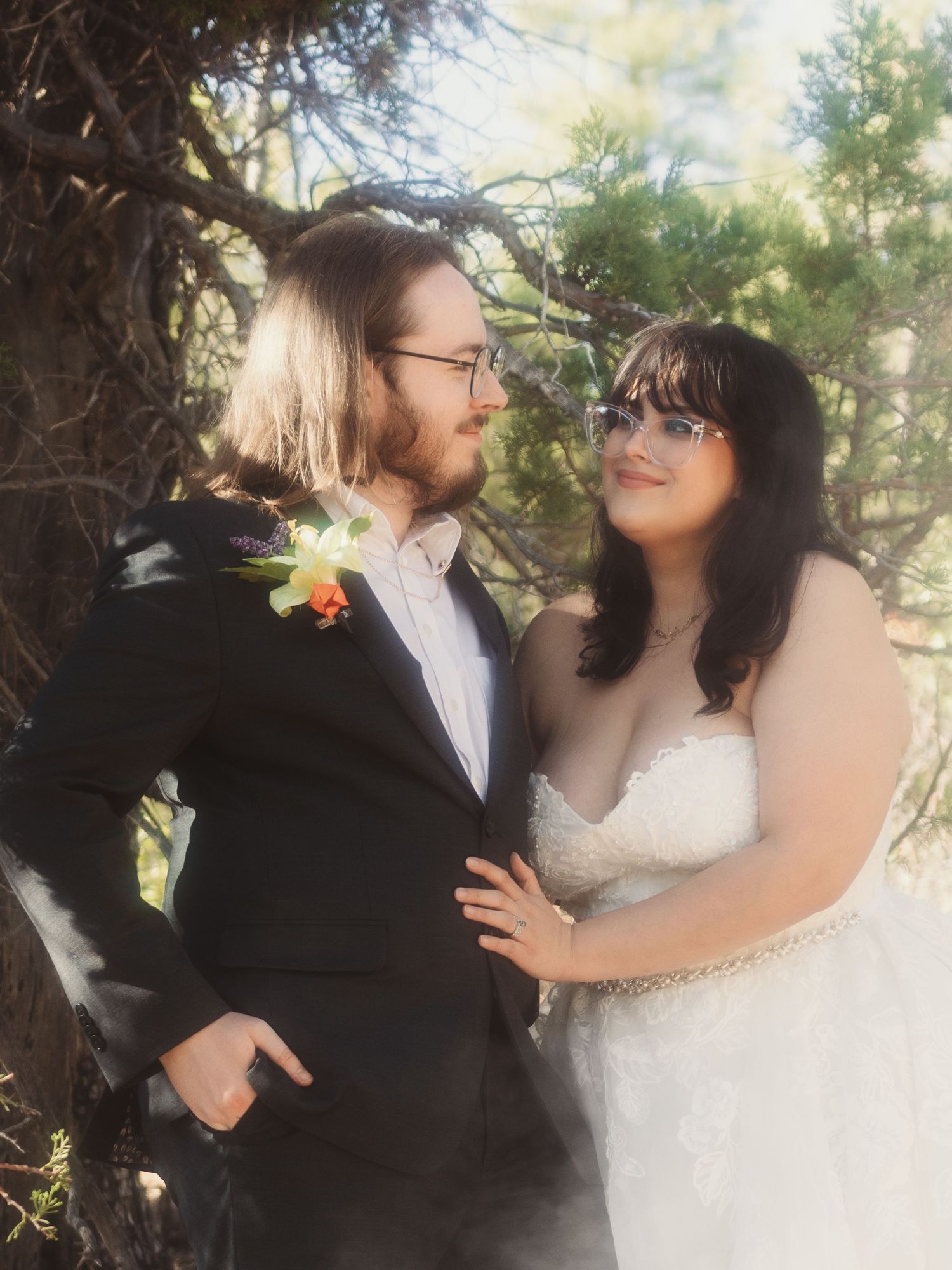 Bride and groom in forest setting share a loving glance among pine trees.