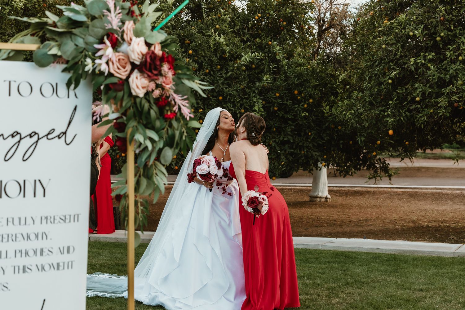 Bride in white gown embracing bridesmaid in red dress near floral arch at outdoor wedding.