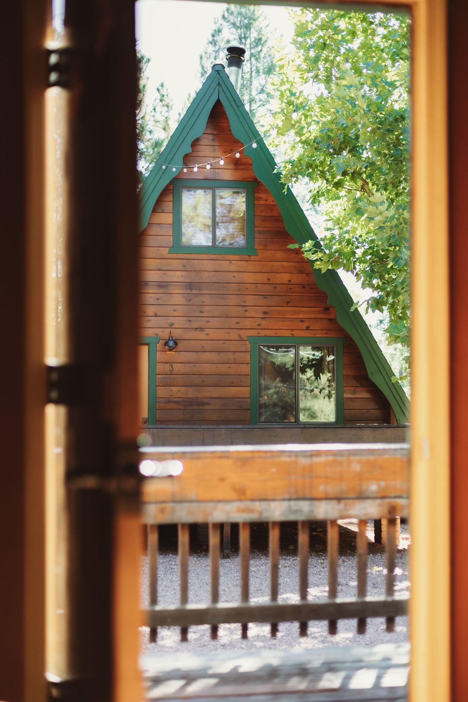 Rustic A-frame wooden cabin with green trim surrounded by trees, viewed through an open door.
