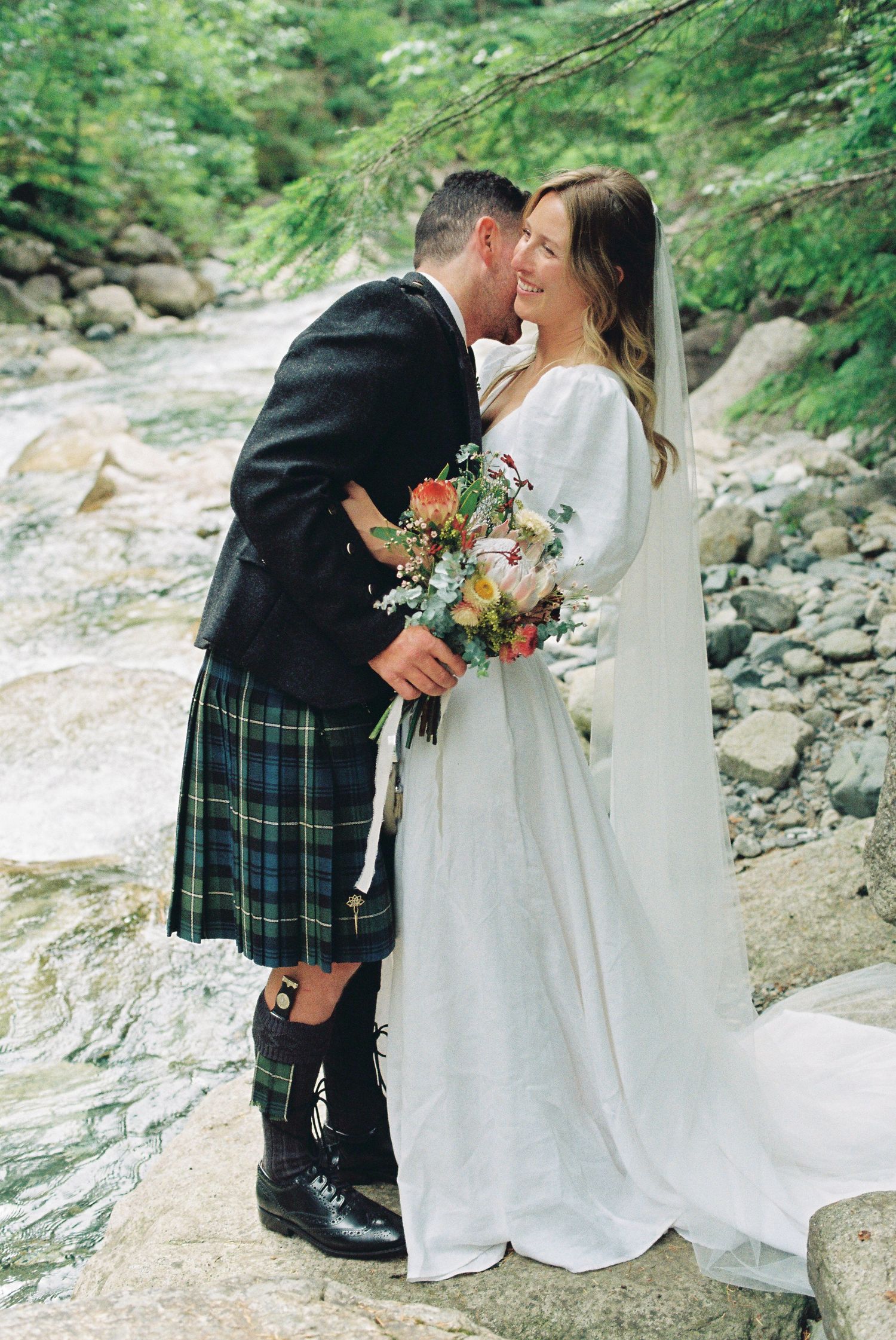 Bride holding colorful bouquet and kilted groom embrace beside rocky mountain stream in lush forest setting.