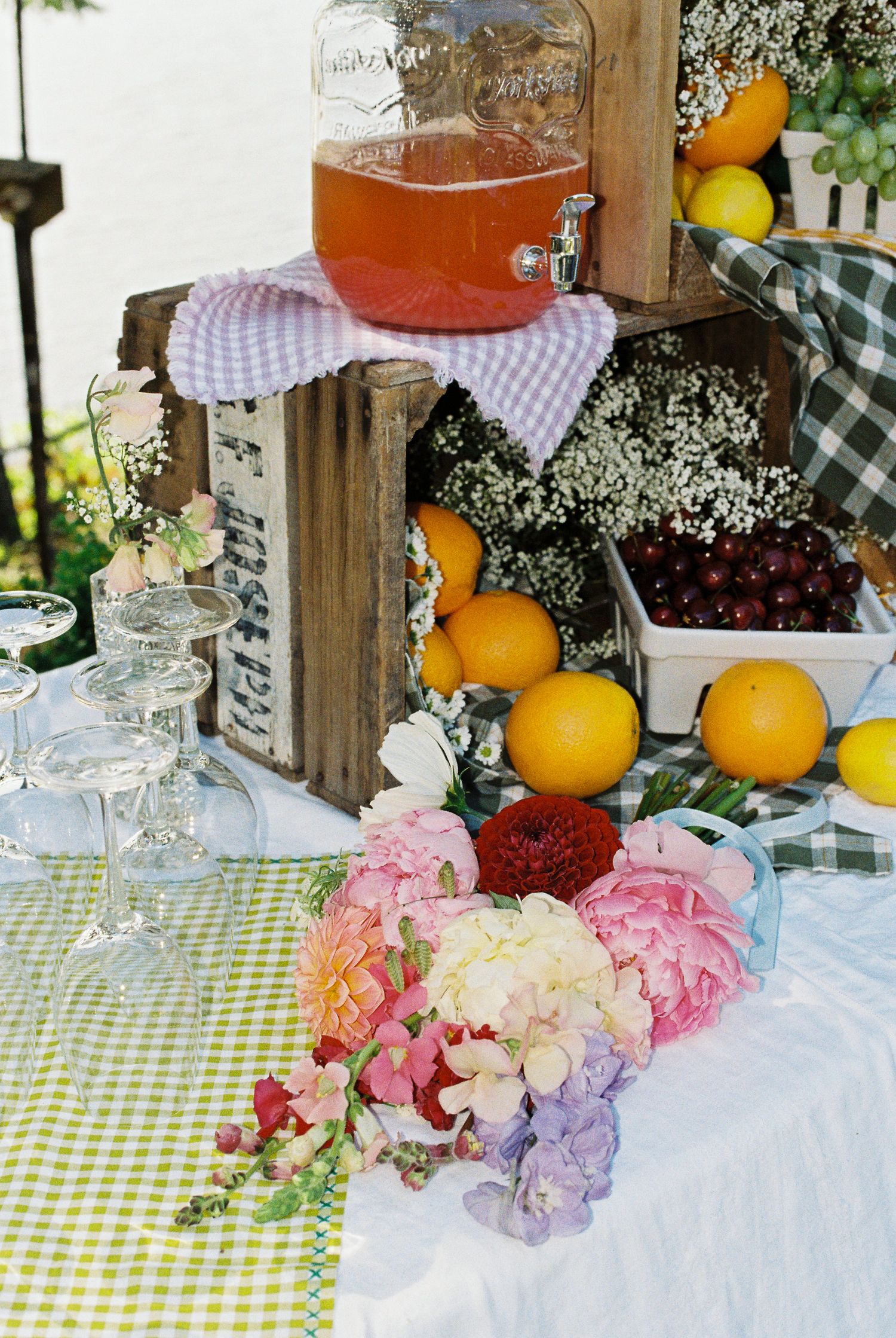 Wedding drink station with pink punch dispenser, fresh fruits, flowers, and glassware on gingham cloth.
