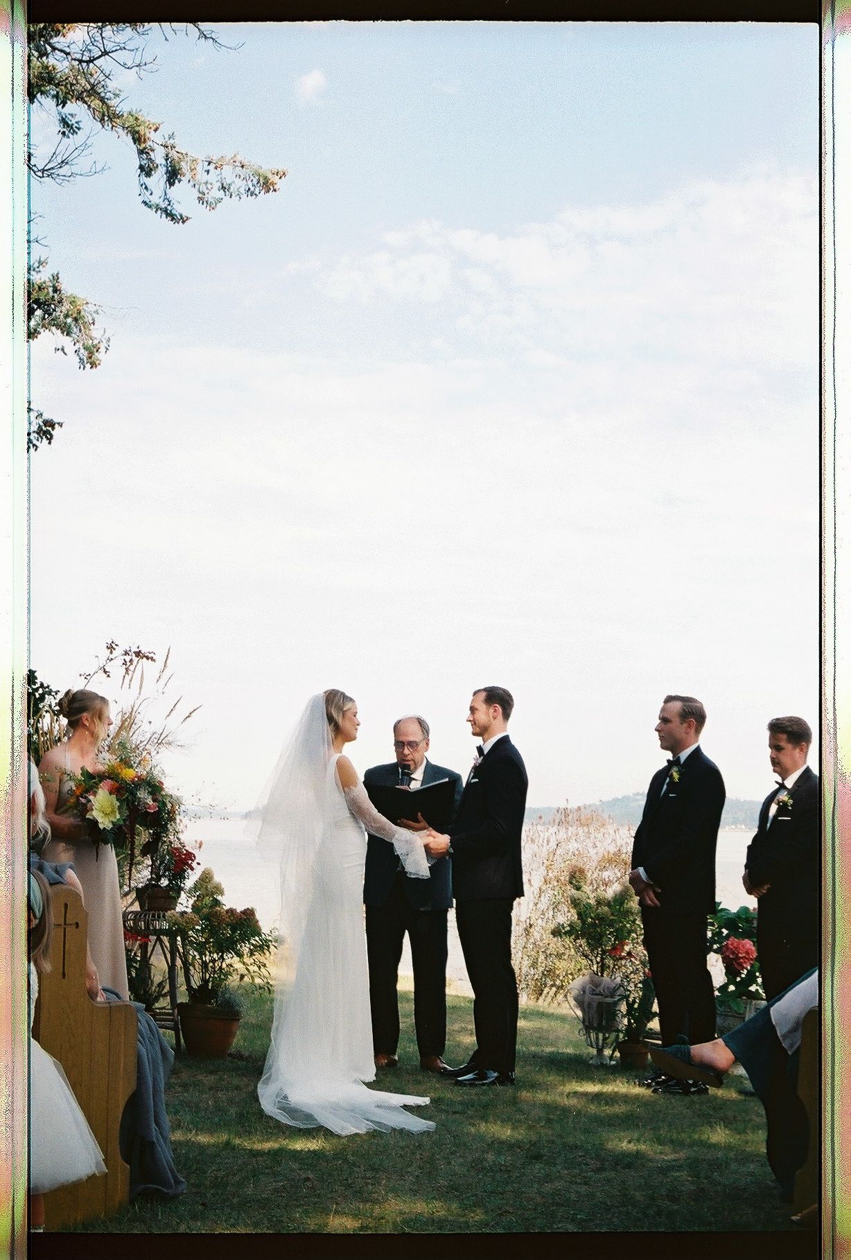 Outdoor waterfront wedding ceremony with bride and groom exchanging vows under open sky.