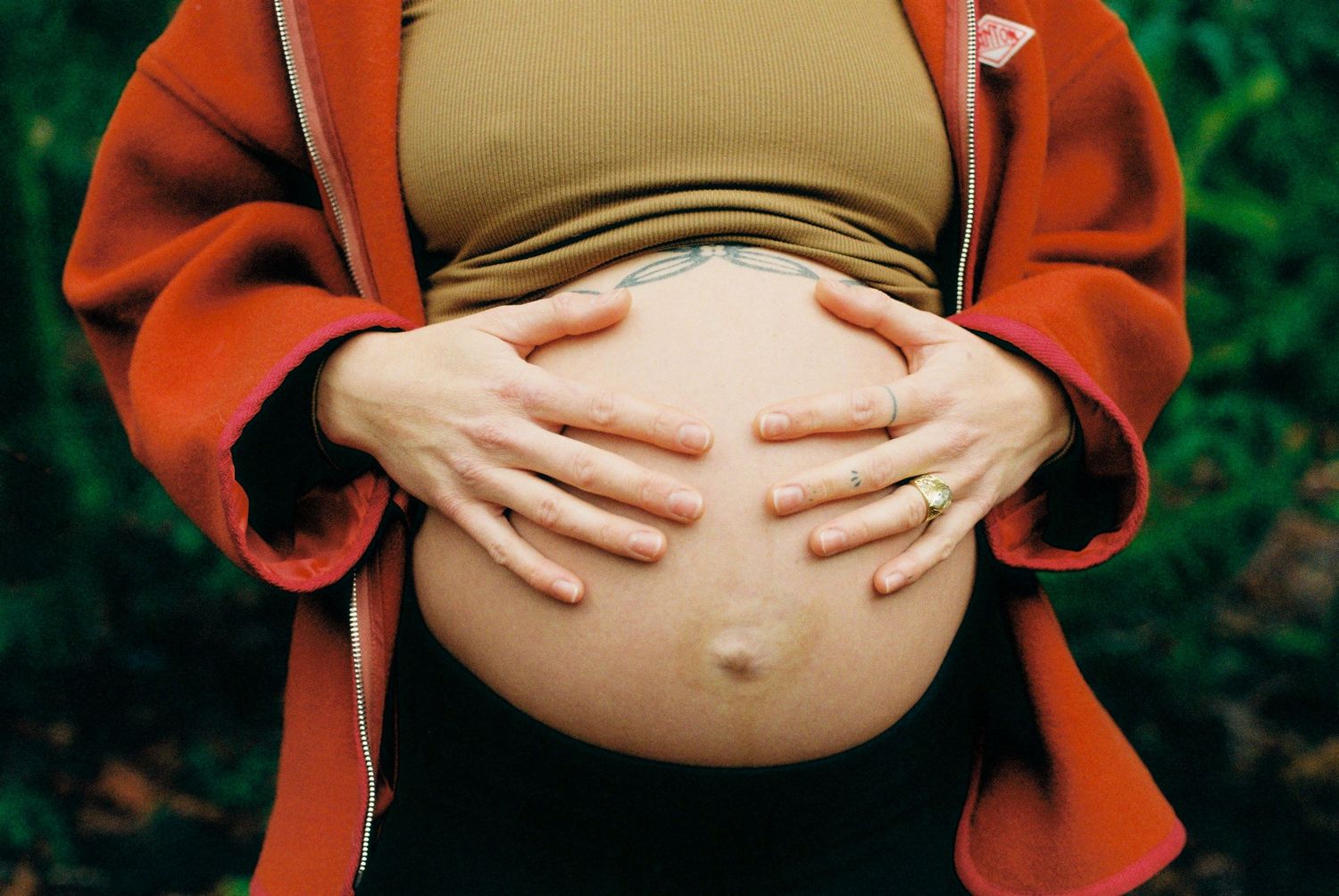 Close-up of pregnant woman in red jacket cradling bare baby bump with both hands.