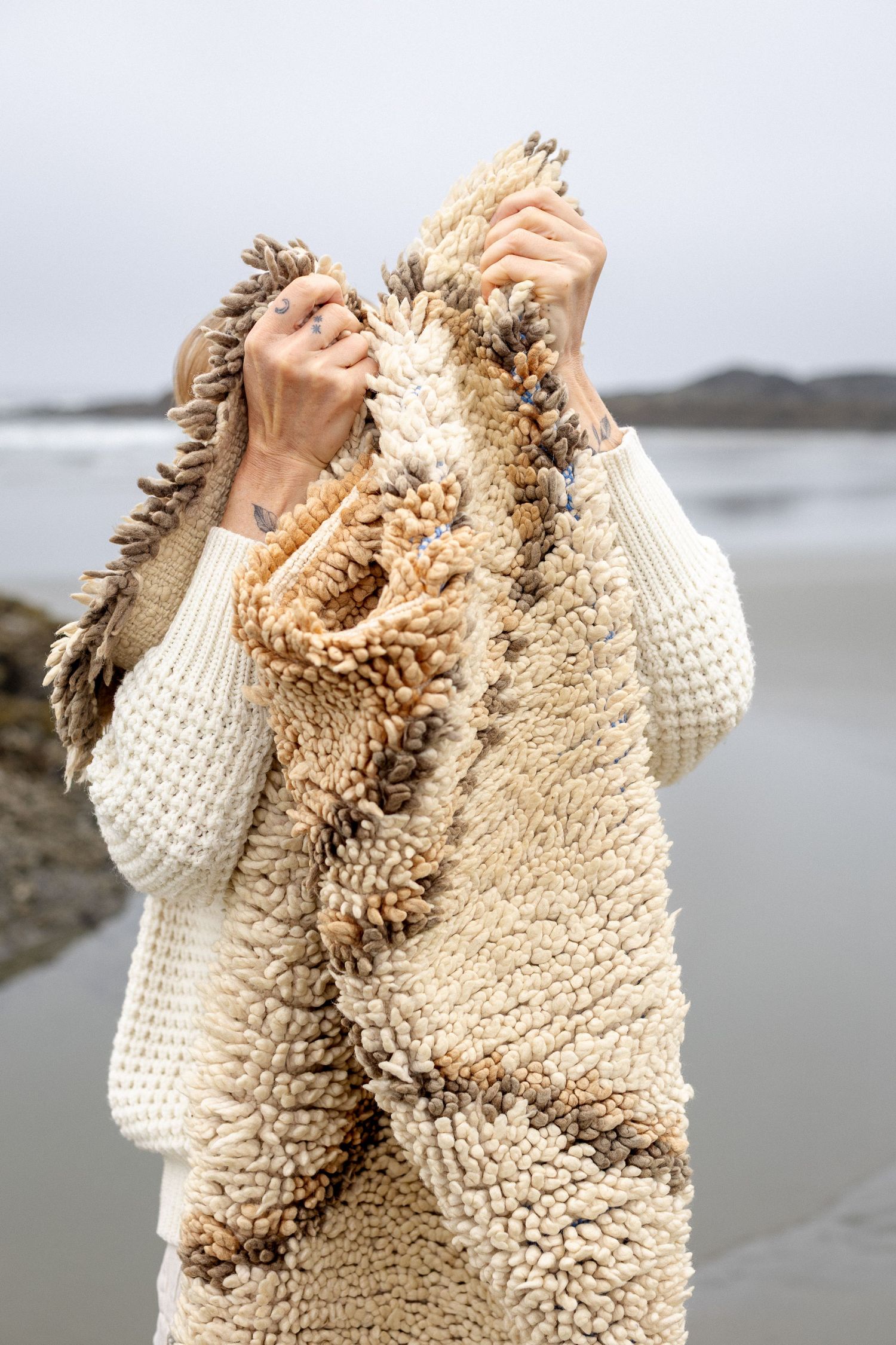 Woman in cream sweater holding a fluffy textured Moroccan rug on a cloudy beach.