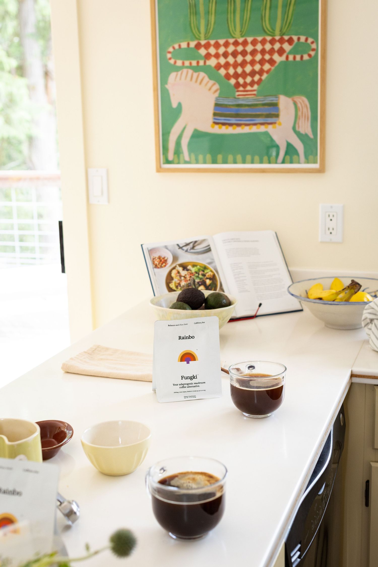 Kitchen counter with black coffee in glass cups, Rainbo product card, avocados, and colorful horse artwork.