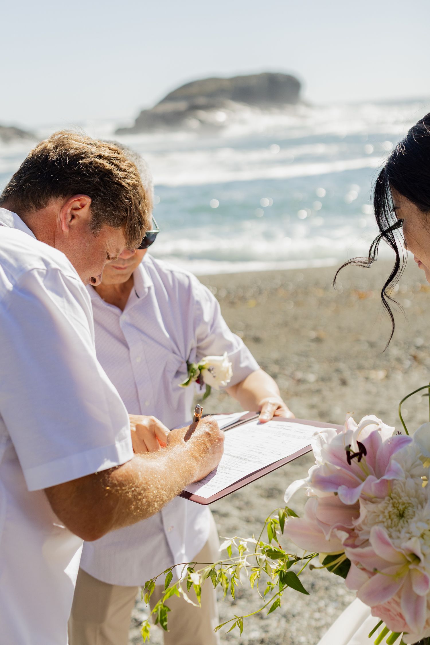 Officiant signs marriage certificate at sunny beach wedding ceremony as bride holds bouquet of pink lilies.