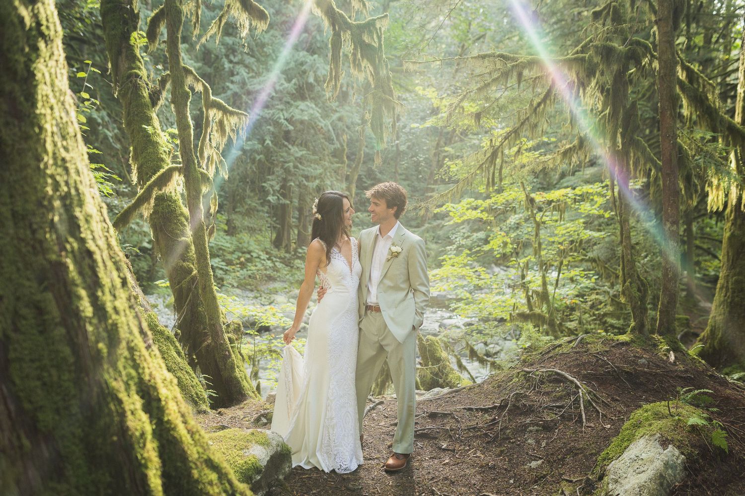 Couple in wedding attire holding hands in a magical sunlit mossy forest with lens flare effect.
