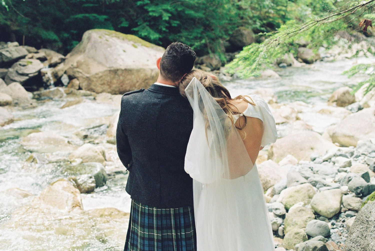 Bride and groom in Scottish kilt stand together gazing at a scenic rocky riverside.