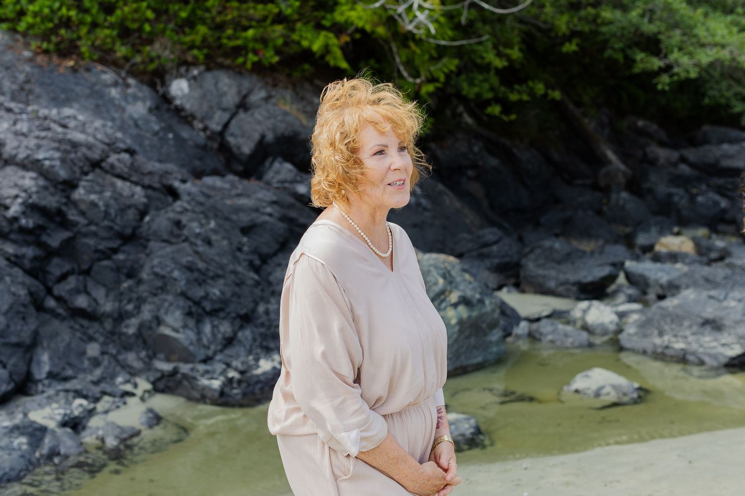 Elegant older woman with curly red hair standing near rocky coastal scenery.