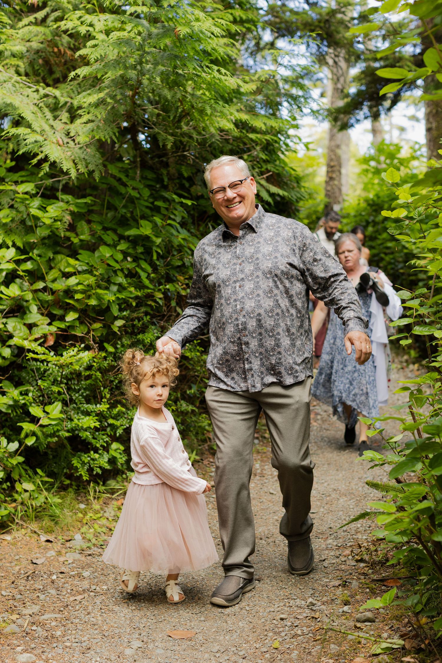 Smiling grandfather walks young flower girl in pink dress along lush green garden path.