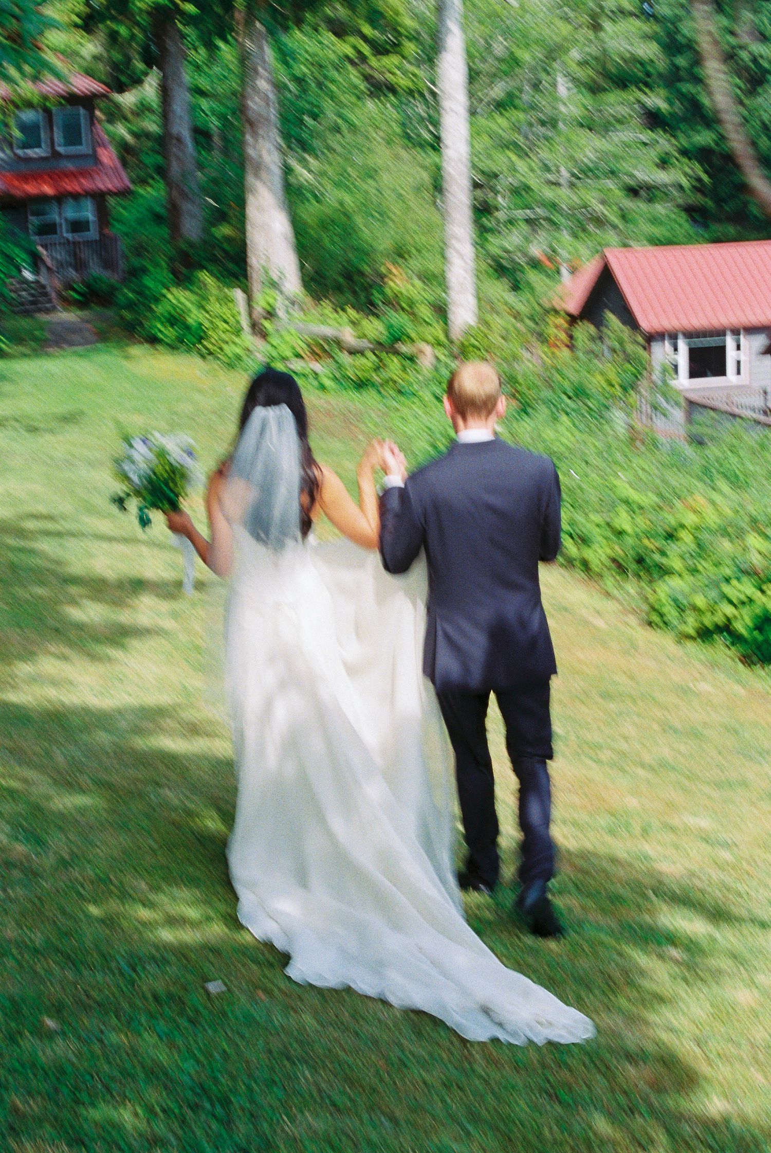 Bride and groom walking hand in hand across green lawn toward rustic cabin in forest.