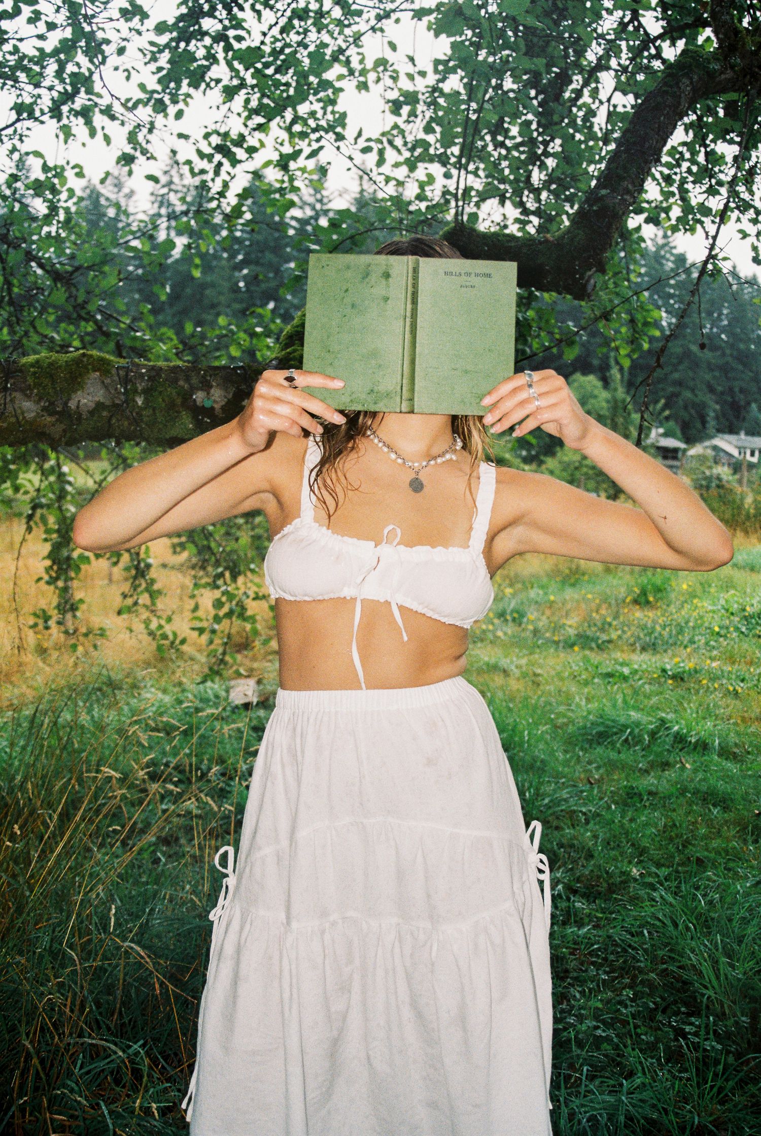 Woman in white bralette and maxi skirt holding a green book over her face outdoors.