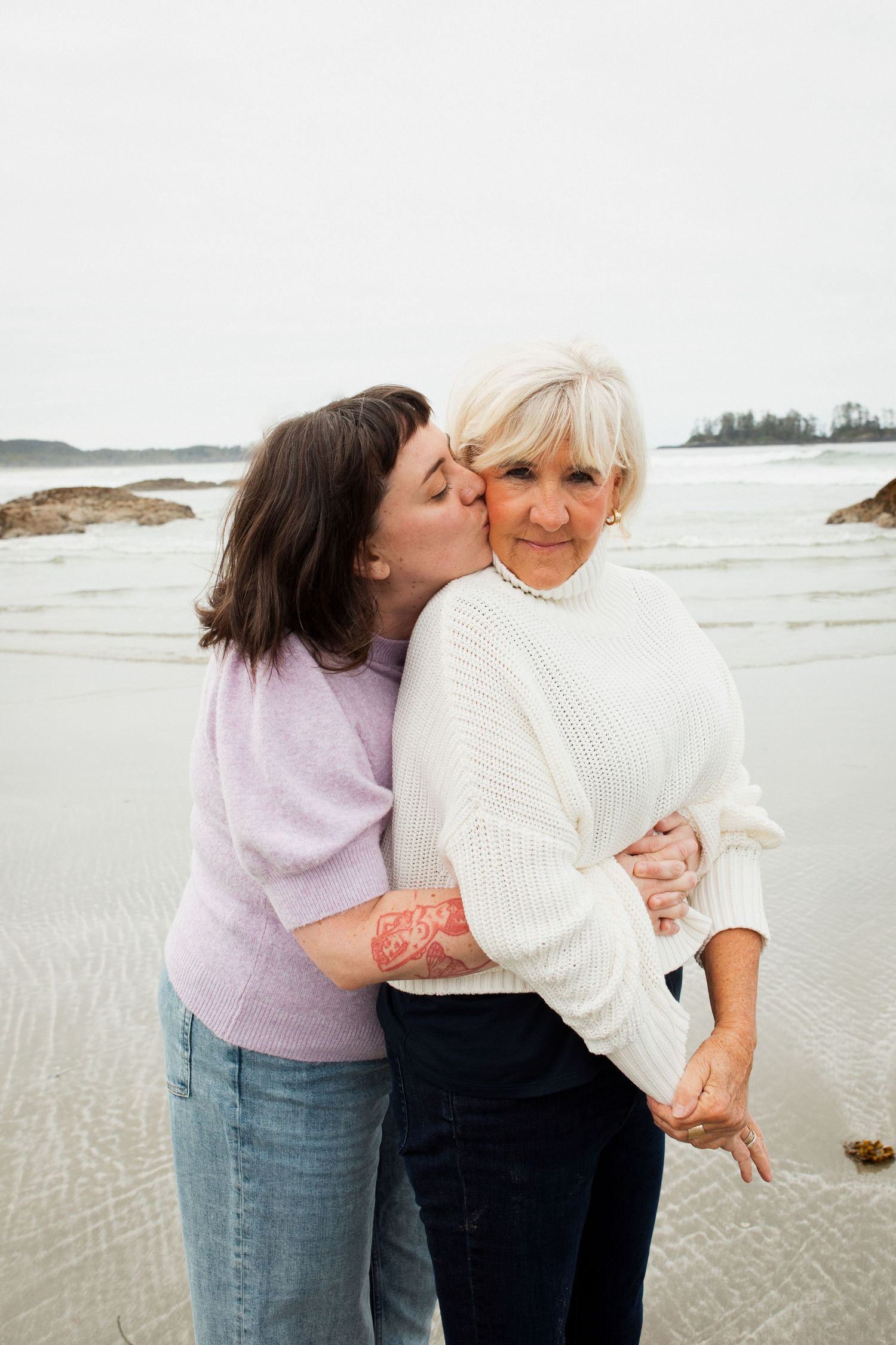 A young woman kisses an older blonde woman on the cheek while standing on a misty beach.
