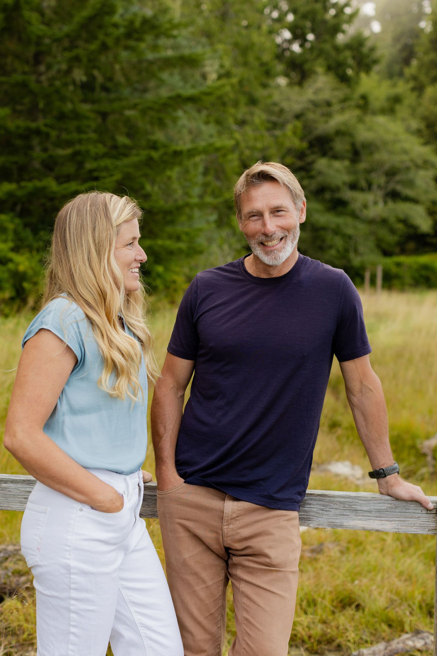 A smiling blonde woman and bearded man lean against a wooden fence in a lush green outdoor setting.