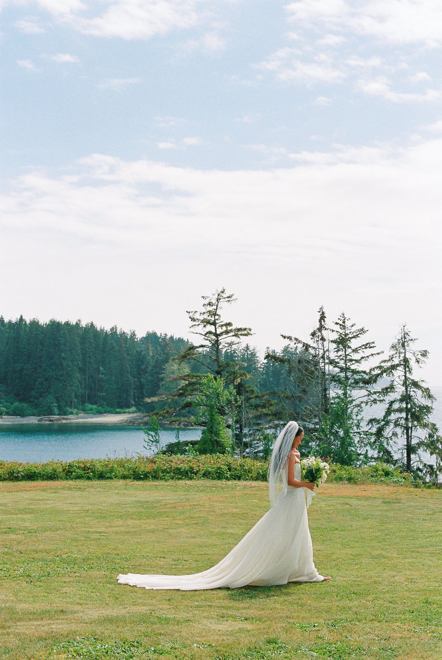 Bride in flowing white gown and veil walks across green lawn with lake and forest backdrop.