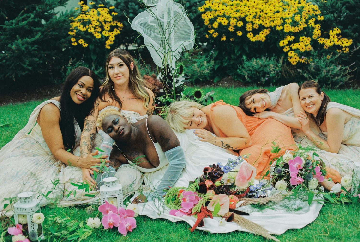 Bride and bridesmaids in colorful dresses lounging on grass surrounded by flowers.