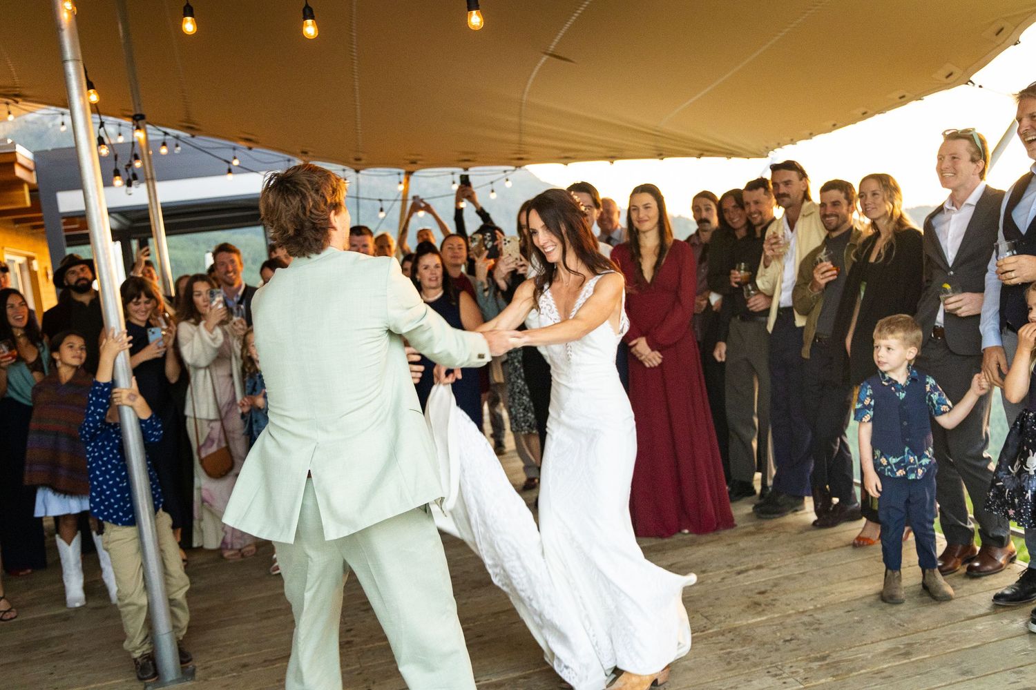 Bride and groom share first dance on wooden floor surrounded by cheering wedding guests under tent.