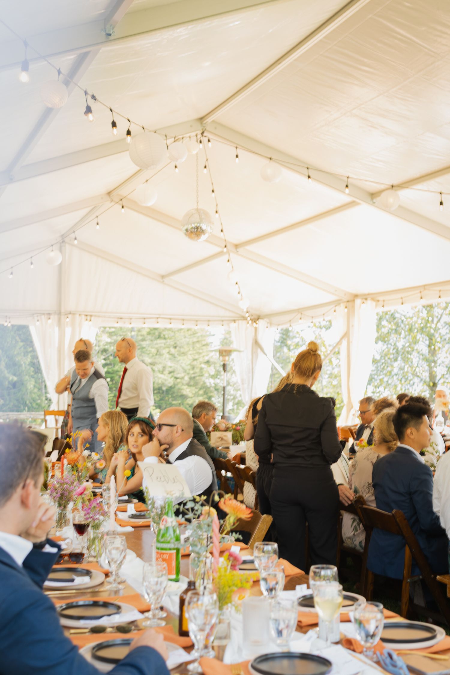 Wedding reception guests dine under a white tent with string lights and floral centerpieces.