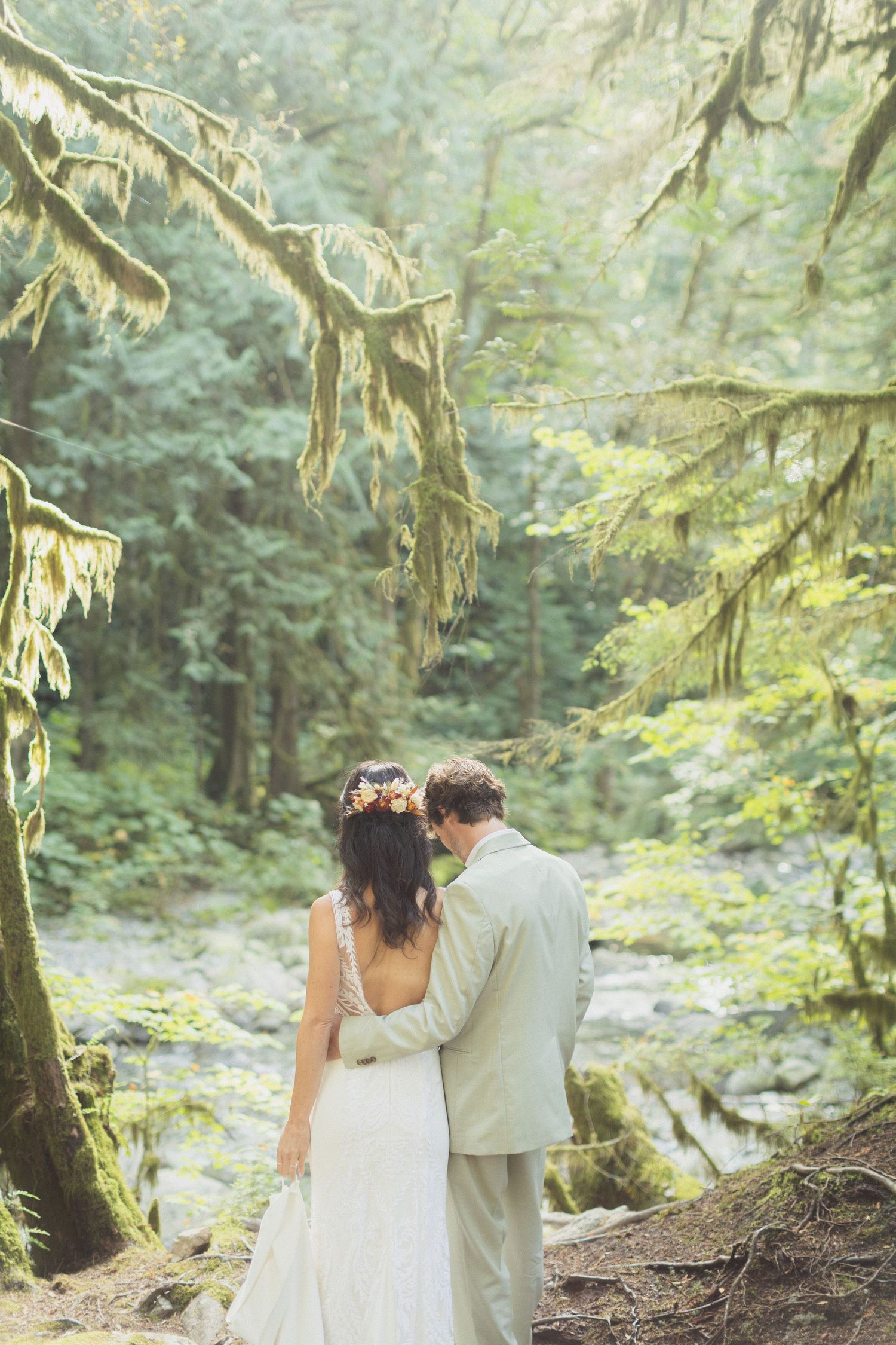 Bride and groom embracing while gazing at a serene forest creek in a moss-covered woodland.