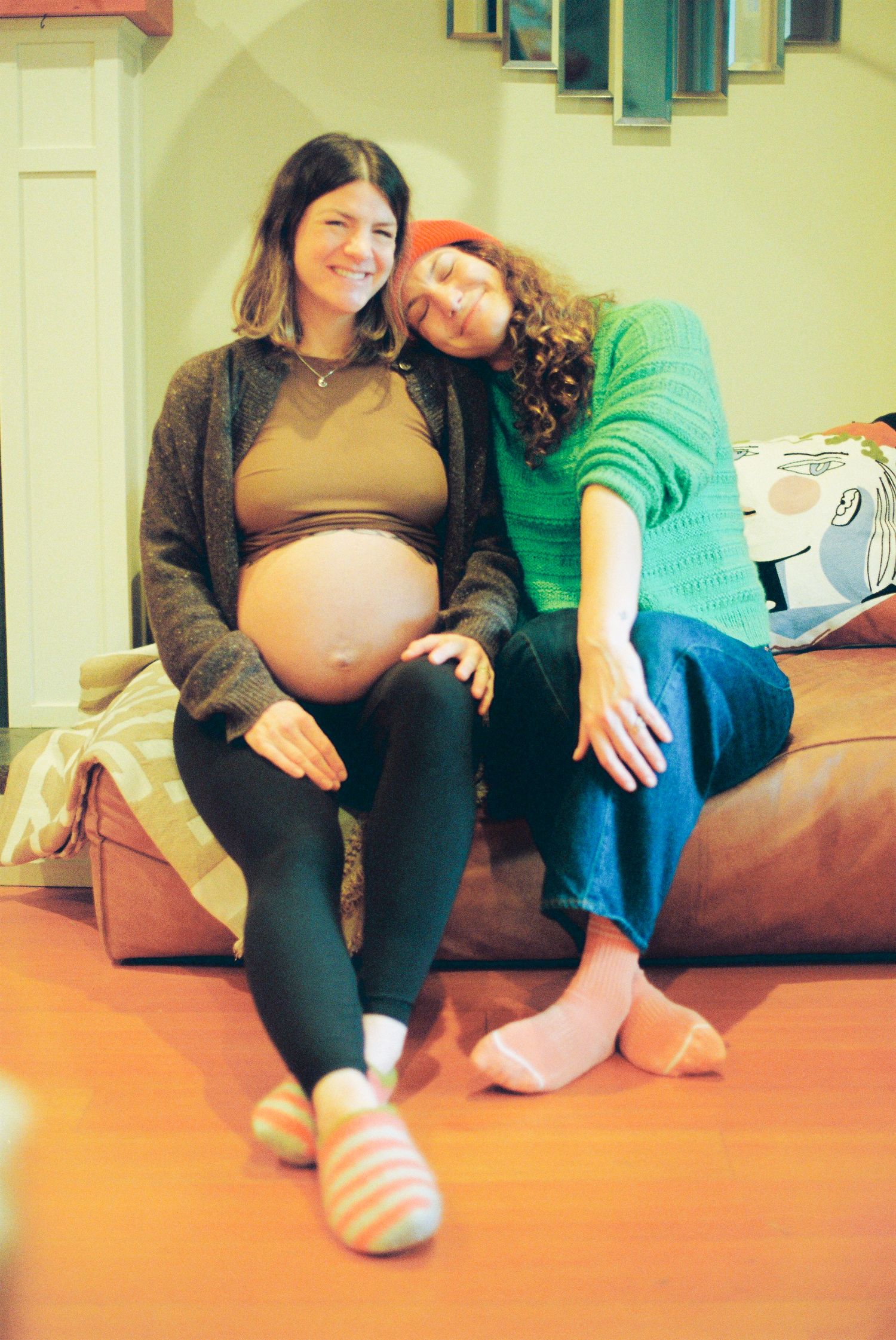 Two smiling women sitting on couch, one pregnant showing baby bump in cozy home.