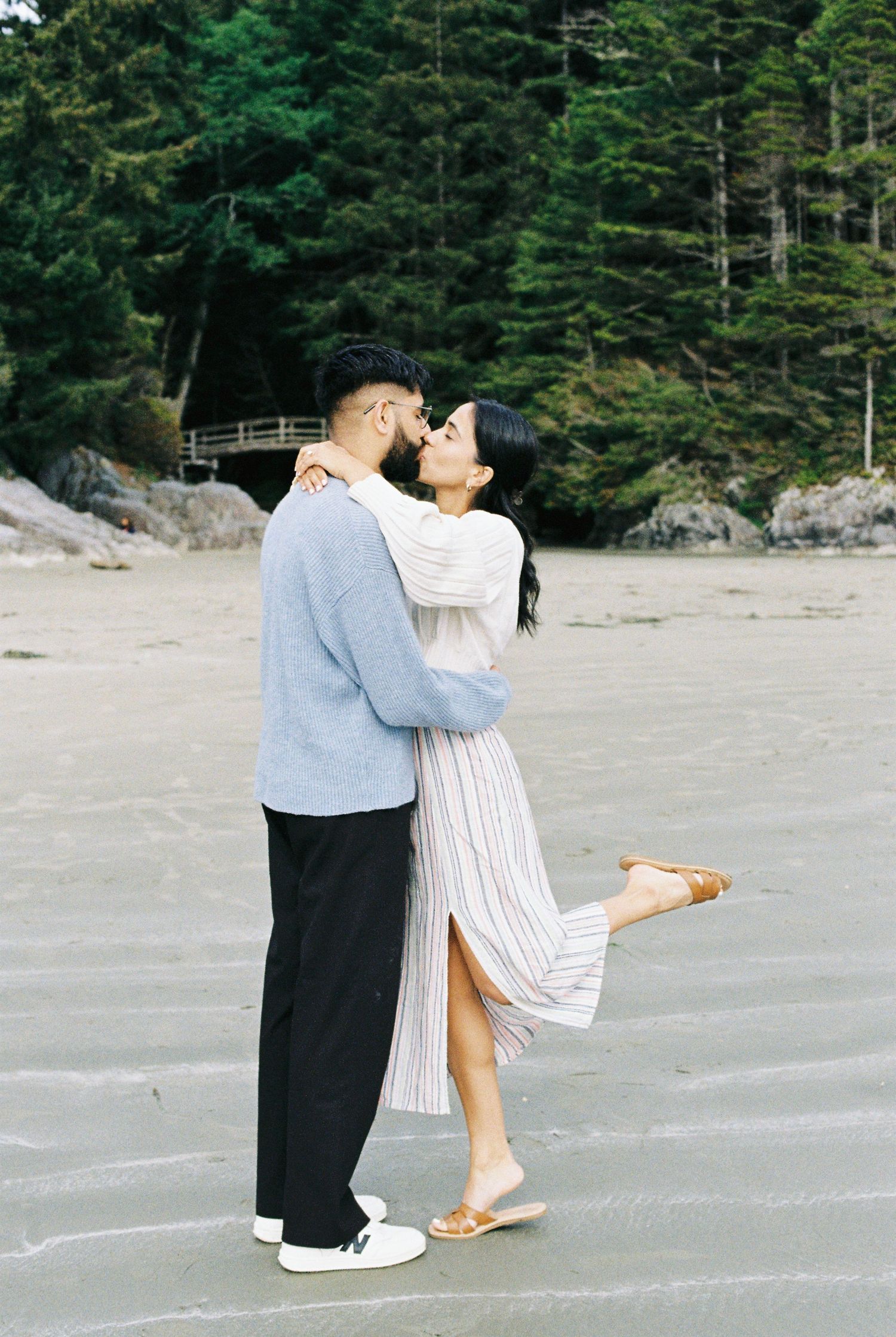 Couple sharing a kiss on a sandy beach surrounded by lush green trees.