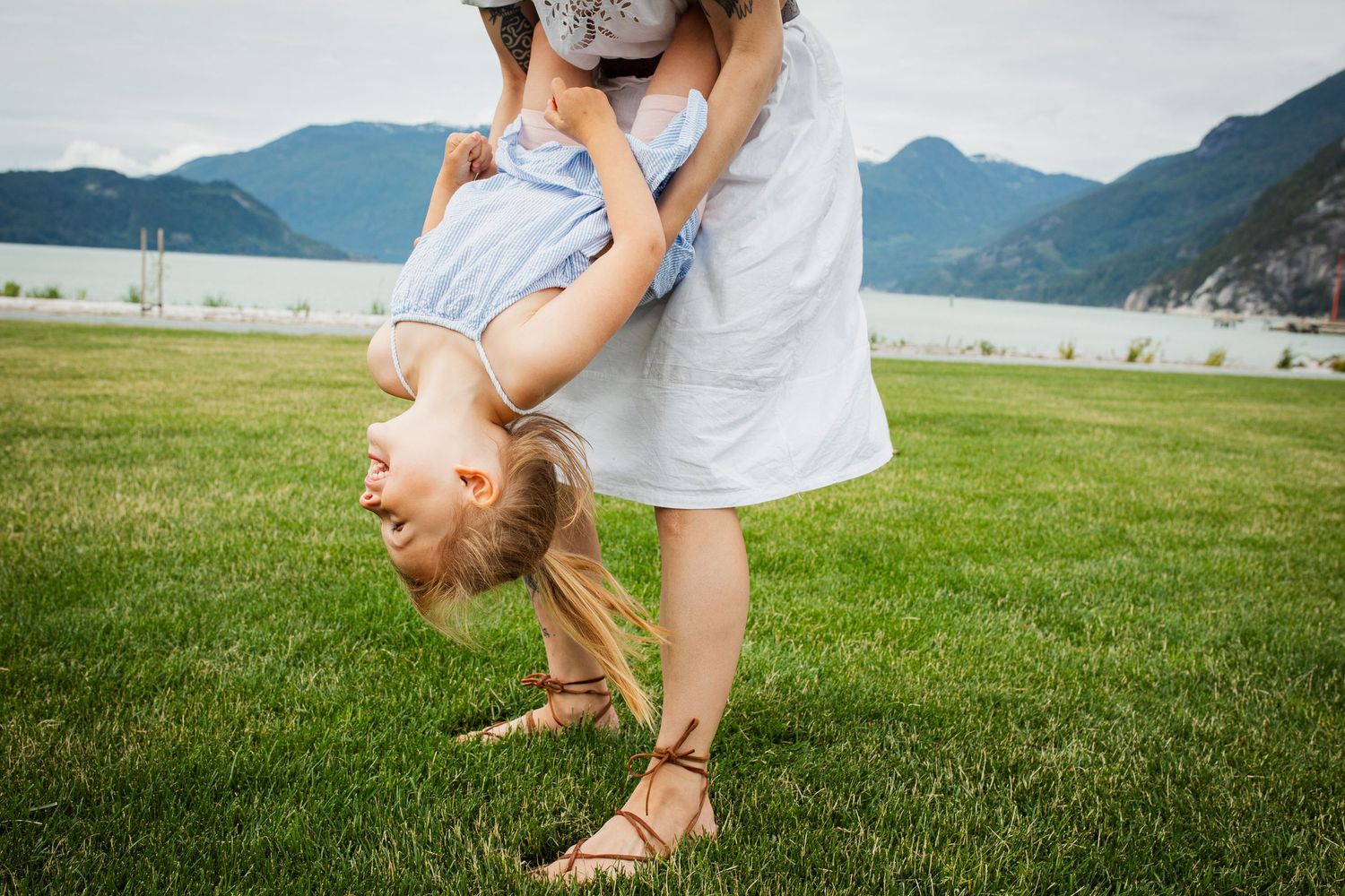 Mother playfully holding young daughter upside down on green lawn with mountain lake in background.