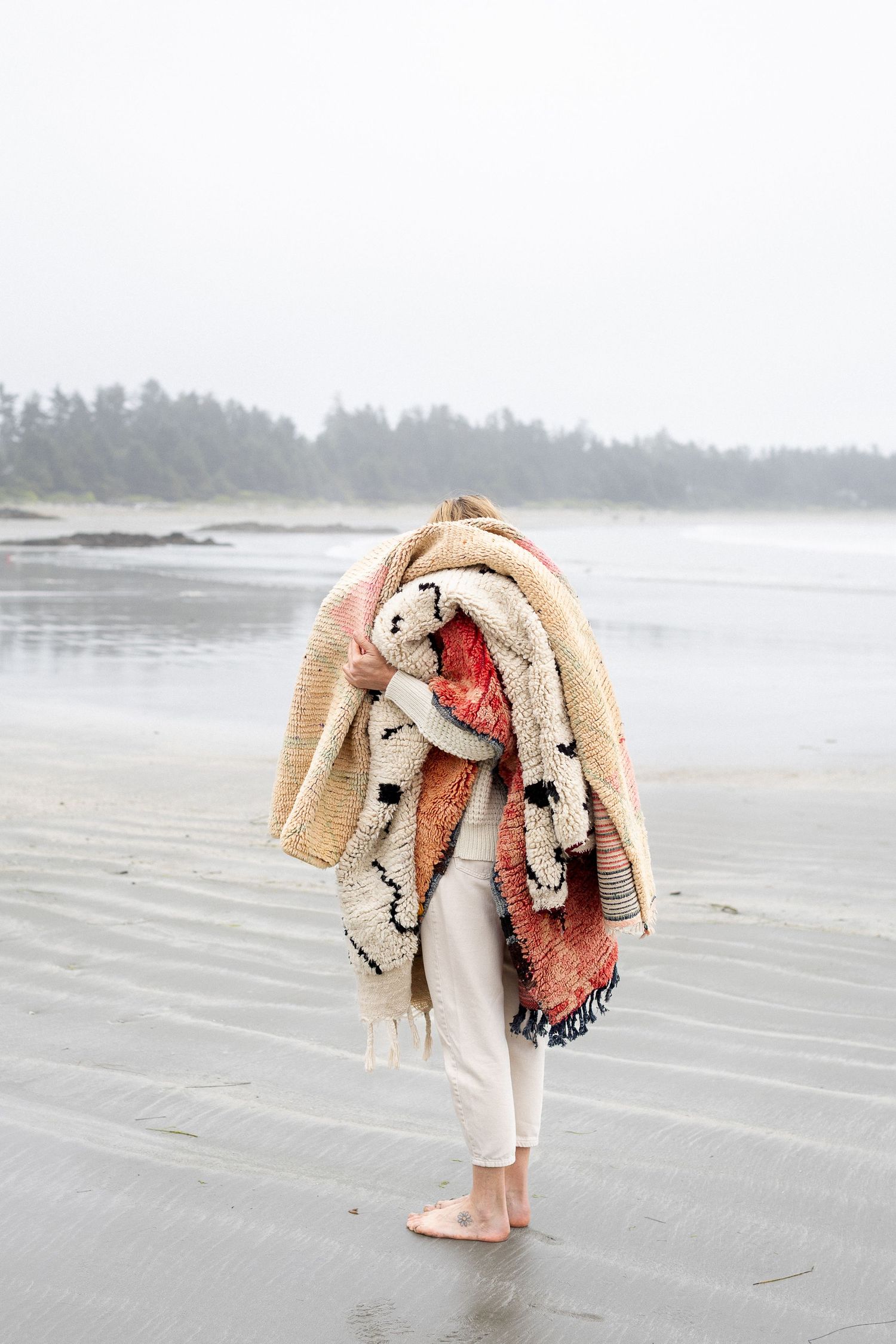 Person carrying colorful layered blankets on a misty beach with dark treeline in background.