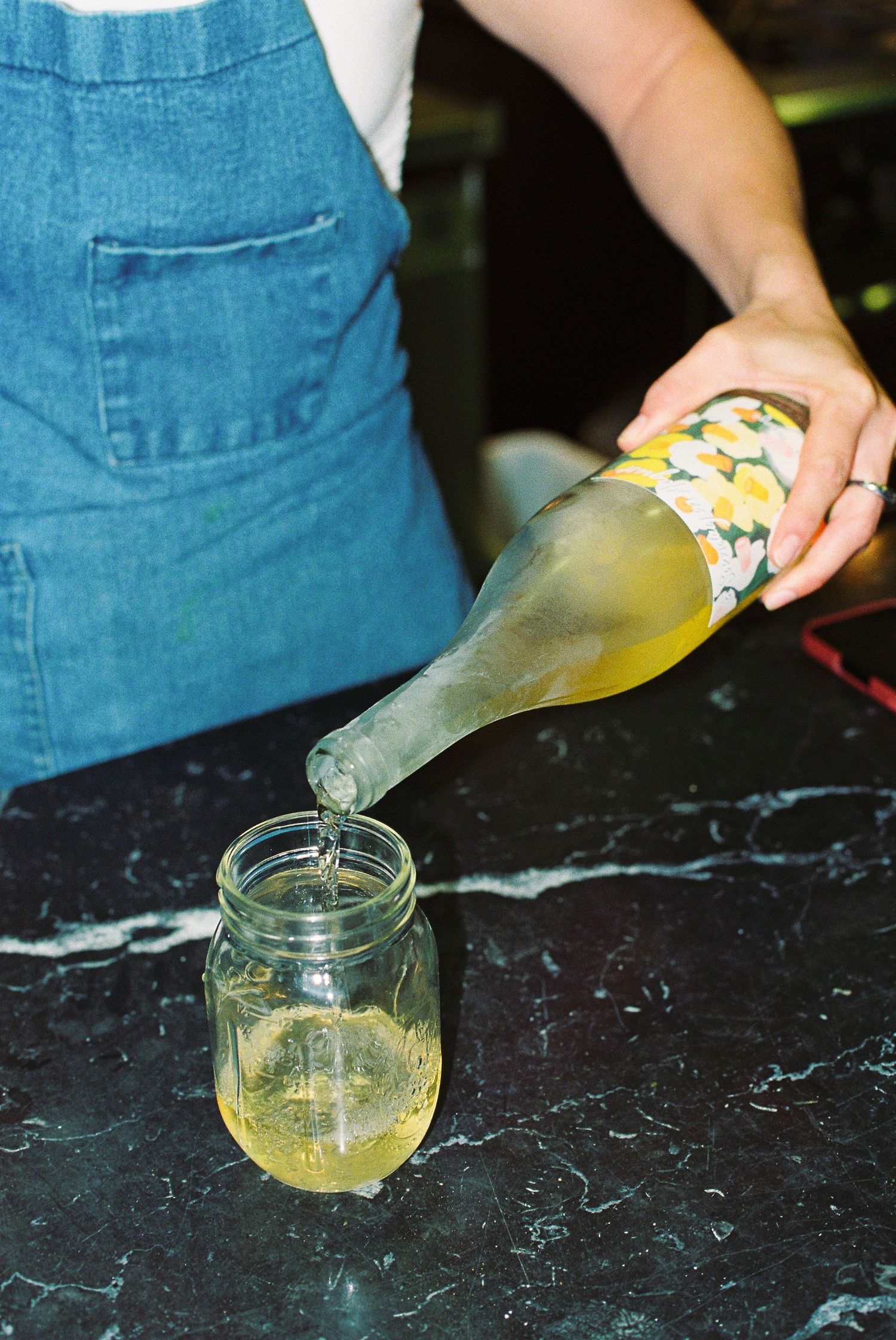 Person in denim apron pouring yellow-green wine from floral-labeled bottle into mason jar on black marble counter.