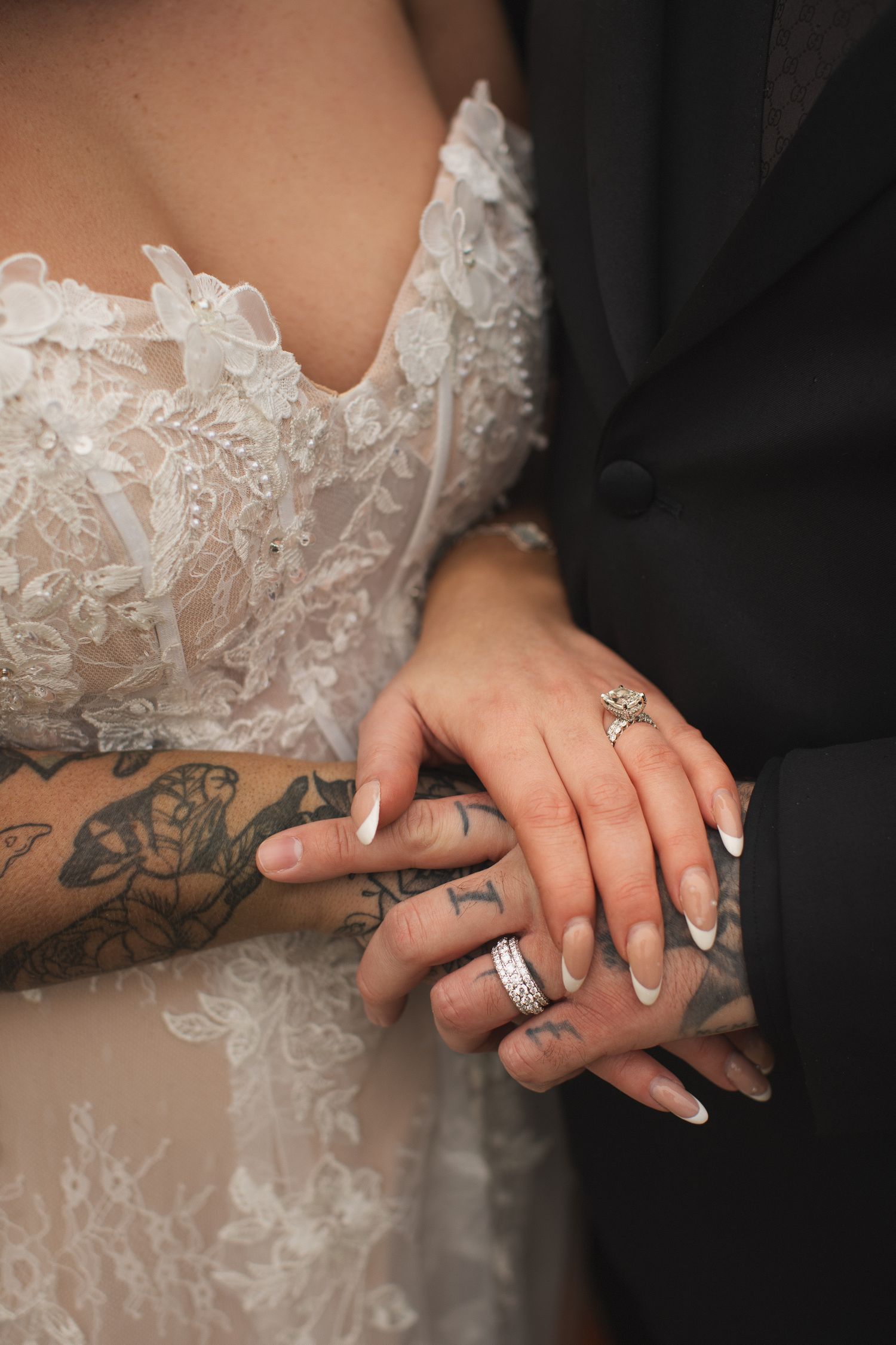 Close-up of tattooed bride and groom holding hands showing wedding rings in lace and suit attire.