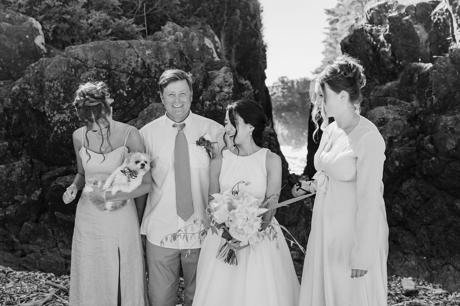 Black and white wedding photo of bride, groom, and bridesmaids with small dog at rocky outdoor ceremony.