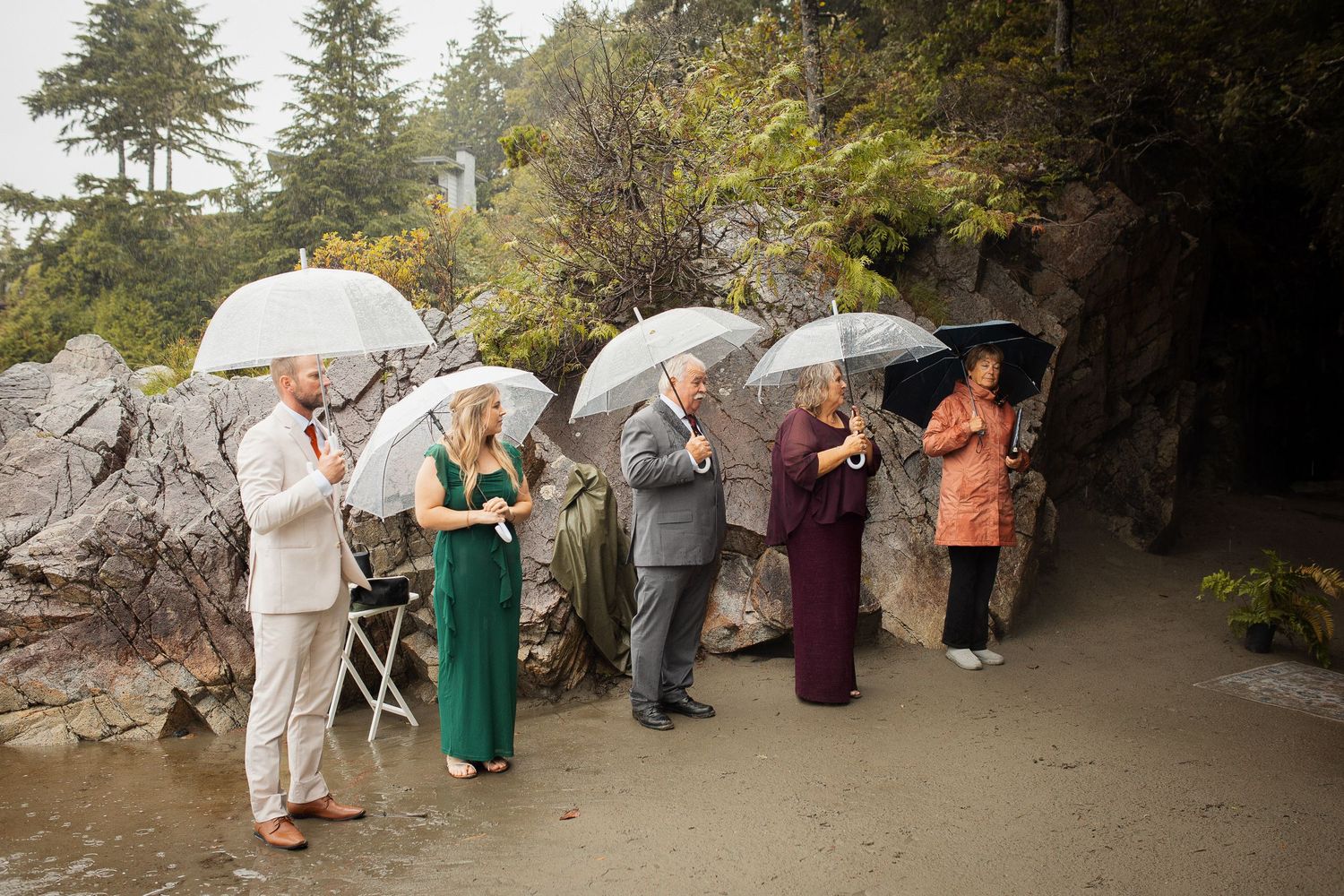 Guests holding clear umbrellas during a rainy outdoor beach wedding ceremony near rocky cliffs.