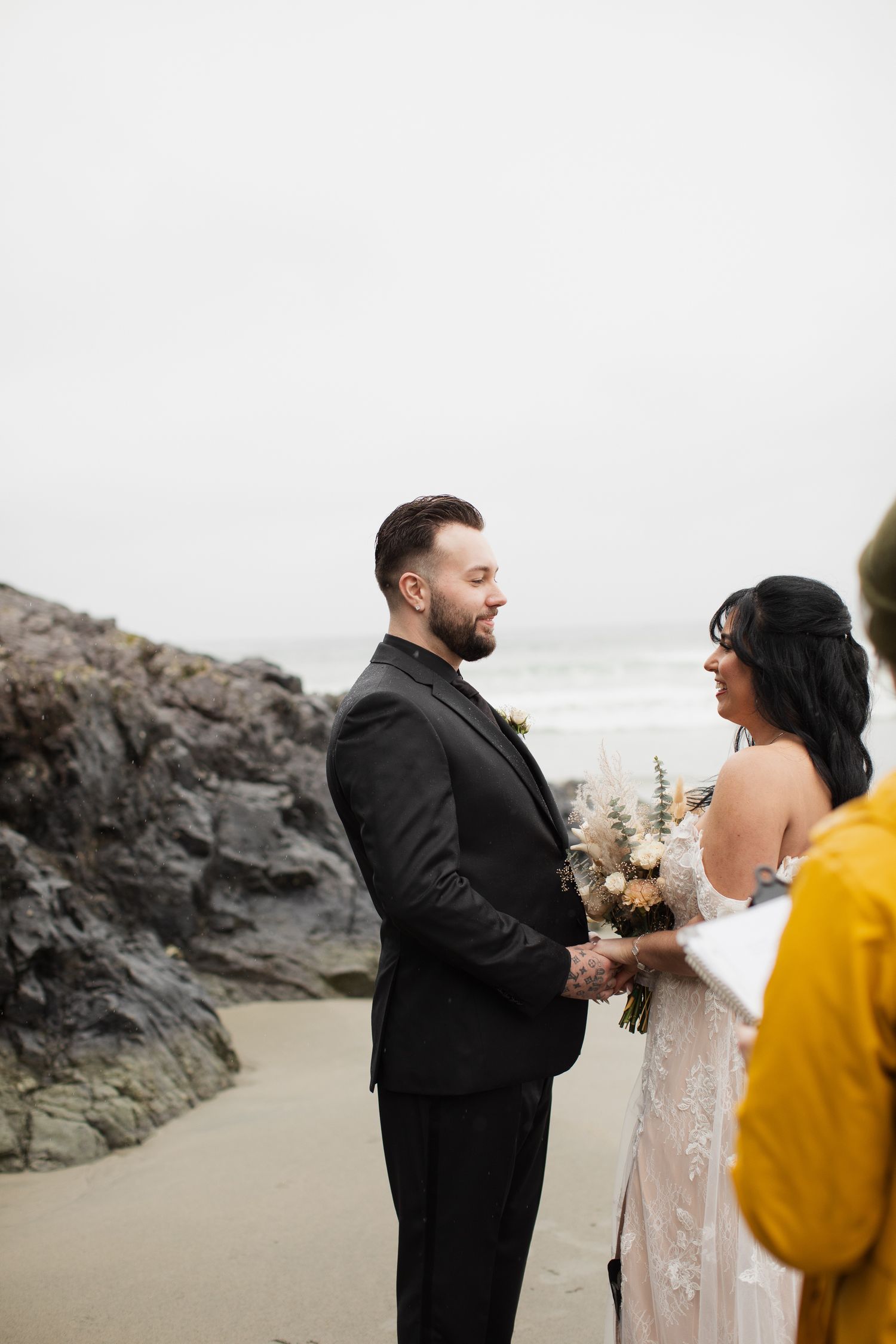 Groom in black suit holds bride's hands during intimate beach wedding ceremony with rocky coastline backdrop.