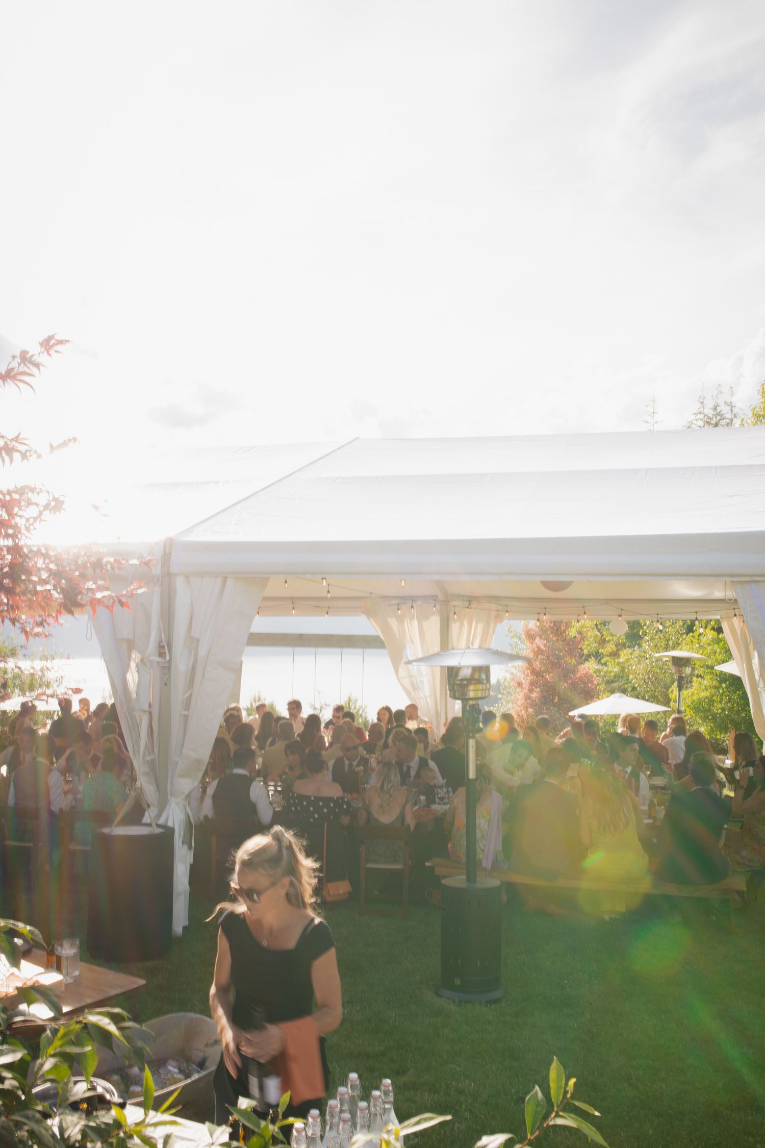 Sunlit outdoor wedding reception under white tent with guests mingling on green lawn.