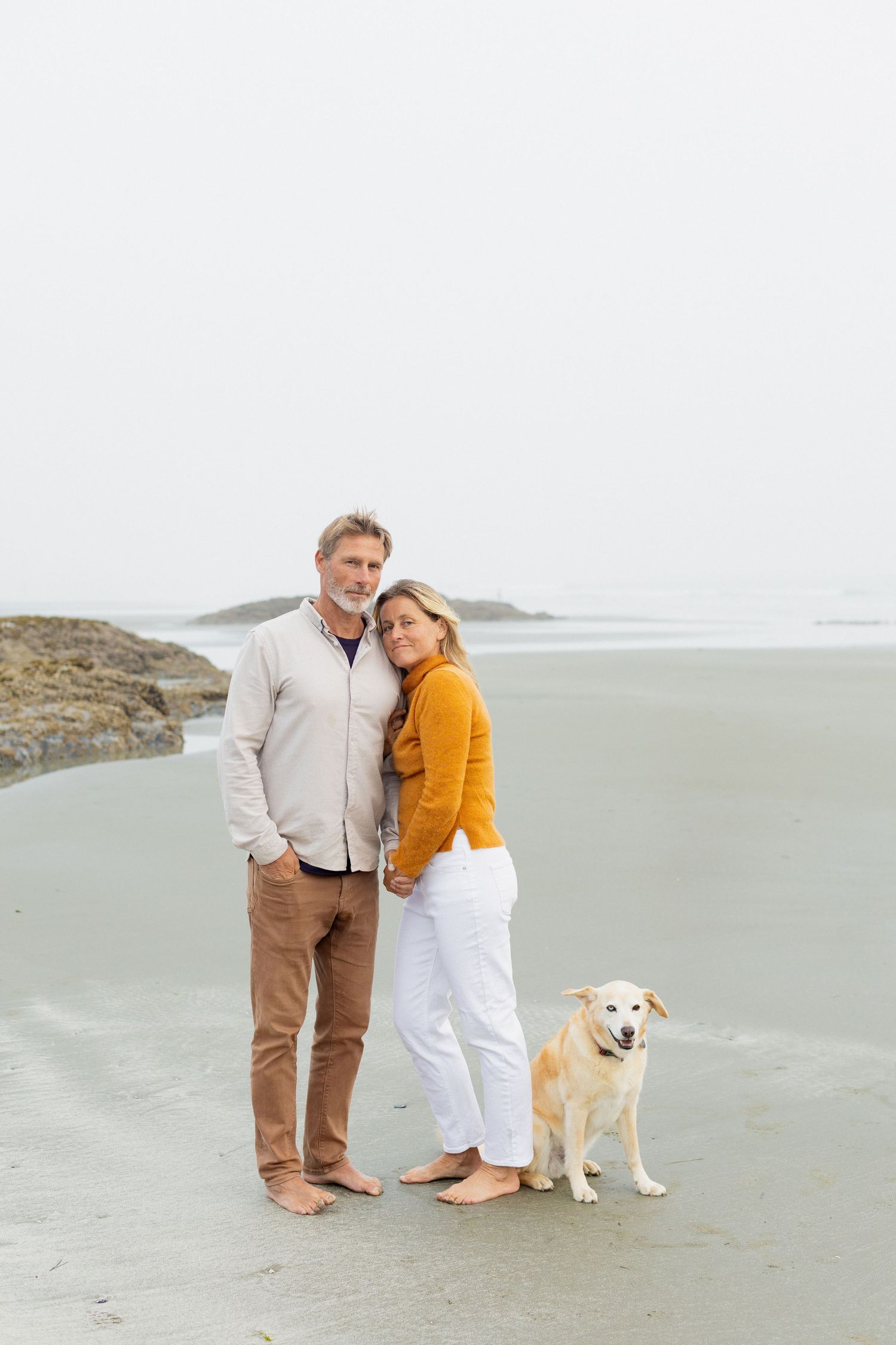 Couple in coordinating outfits standing with golden retriever dog on calm misty beach.