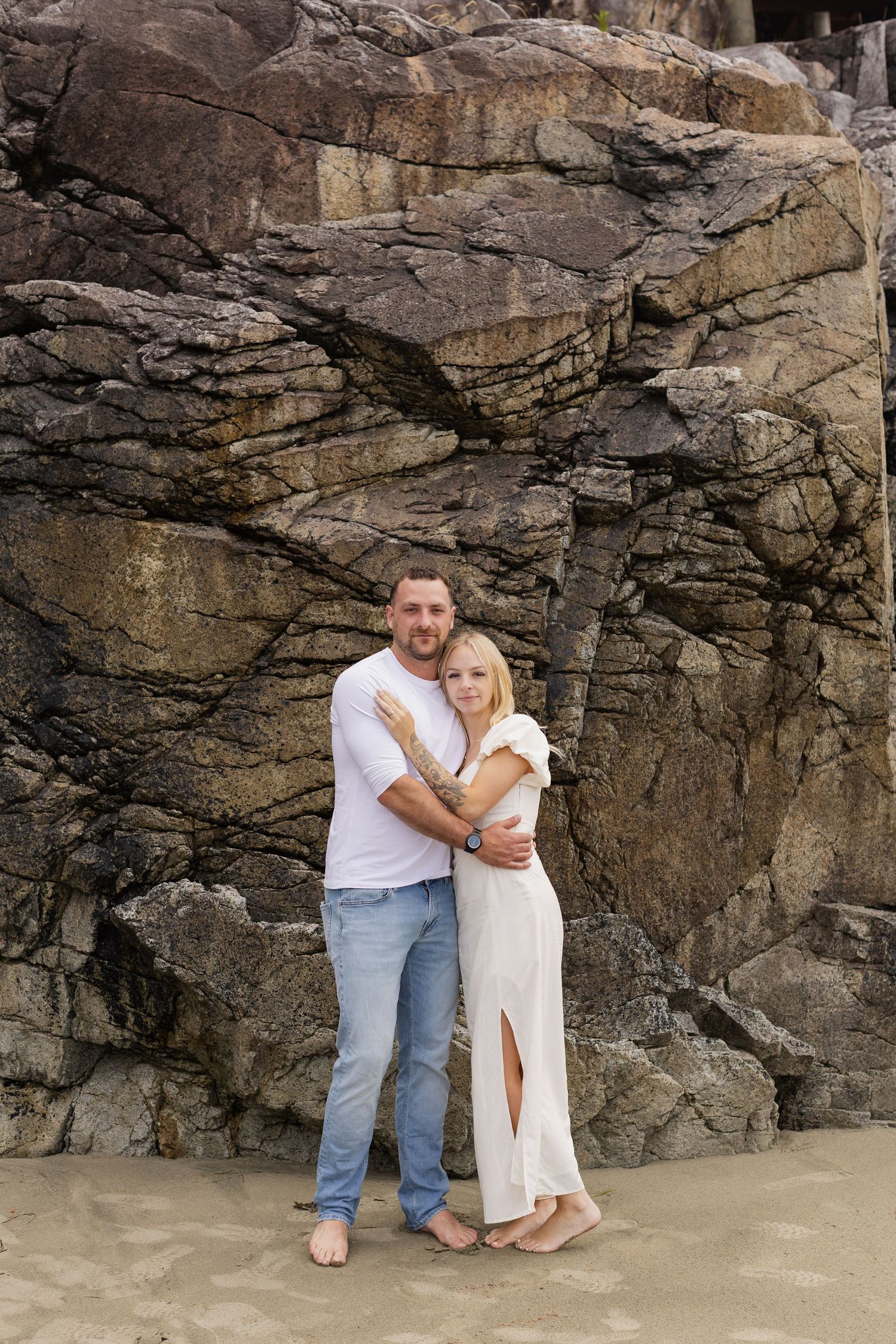 Couple hugging barefoot on sandy beach in front of large rocky cliff formation.