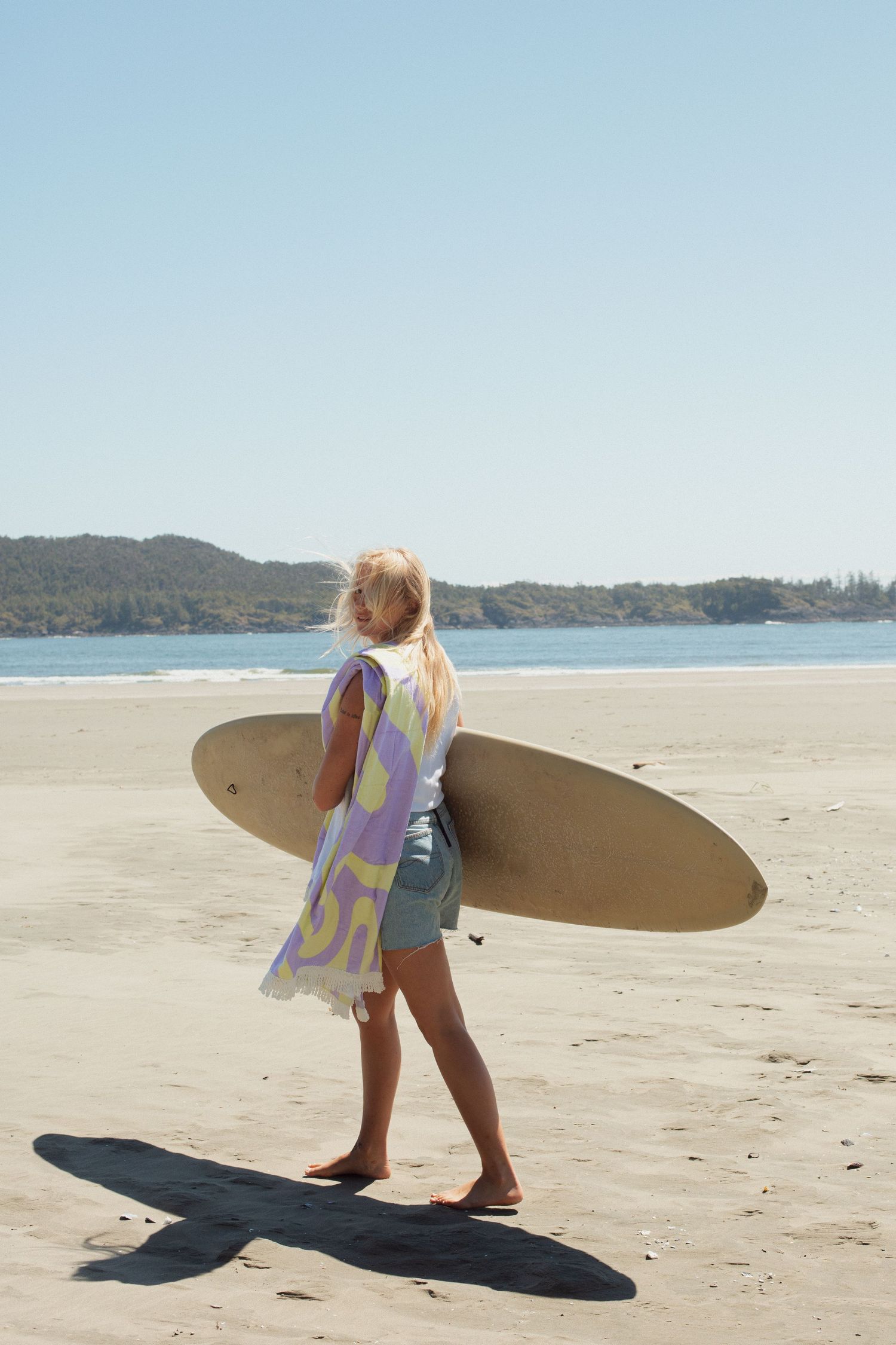 Blonde woman carrying a longboard surfboard on a sunny sandy beach with ocean views.