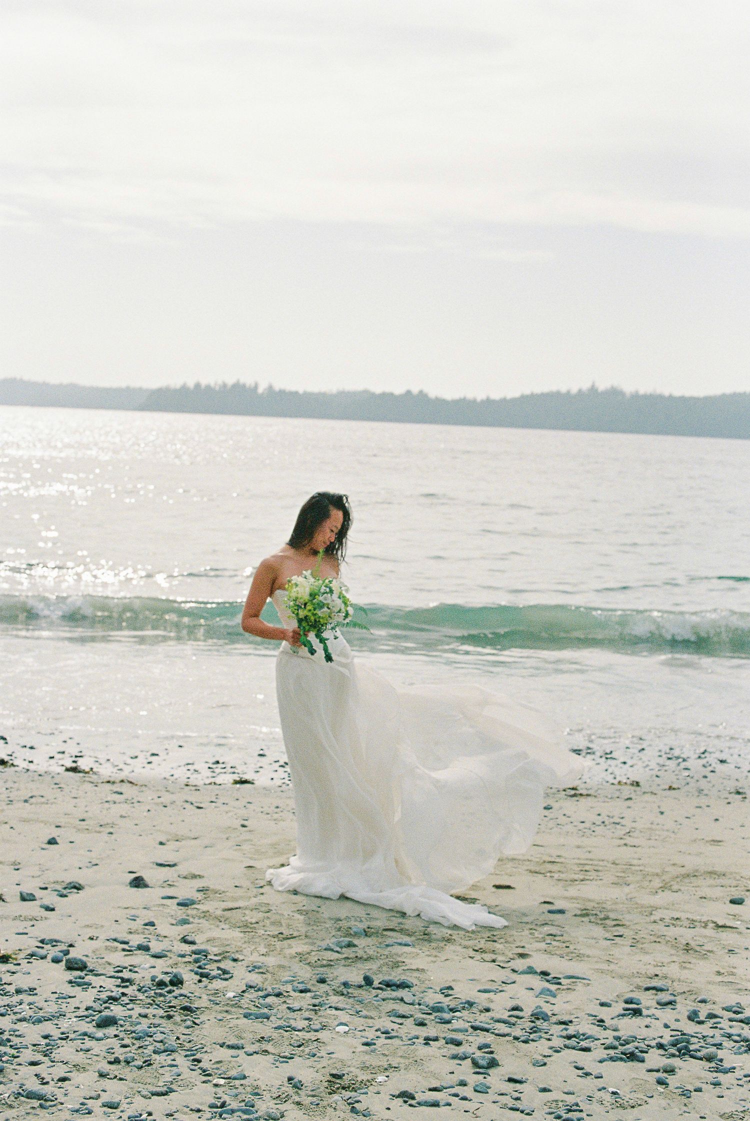 Bride in flowing white gown standing at ocean's edge holding a green bouquet as dress billows in wind.