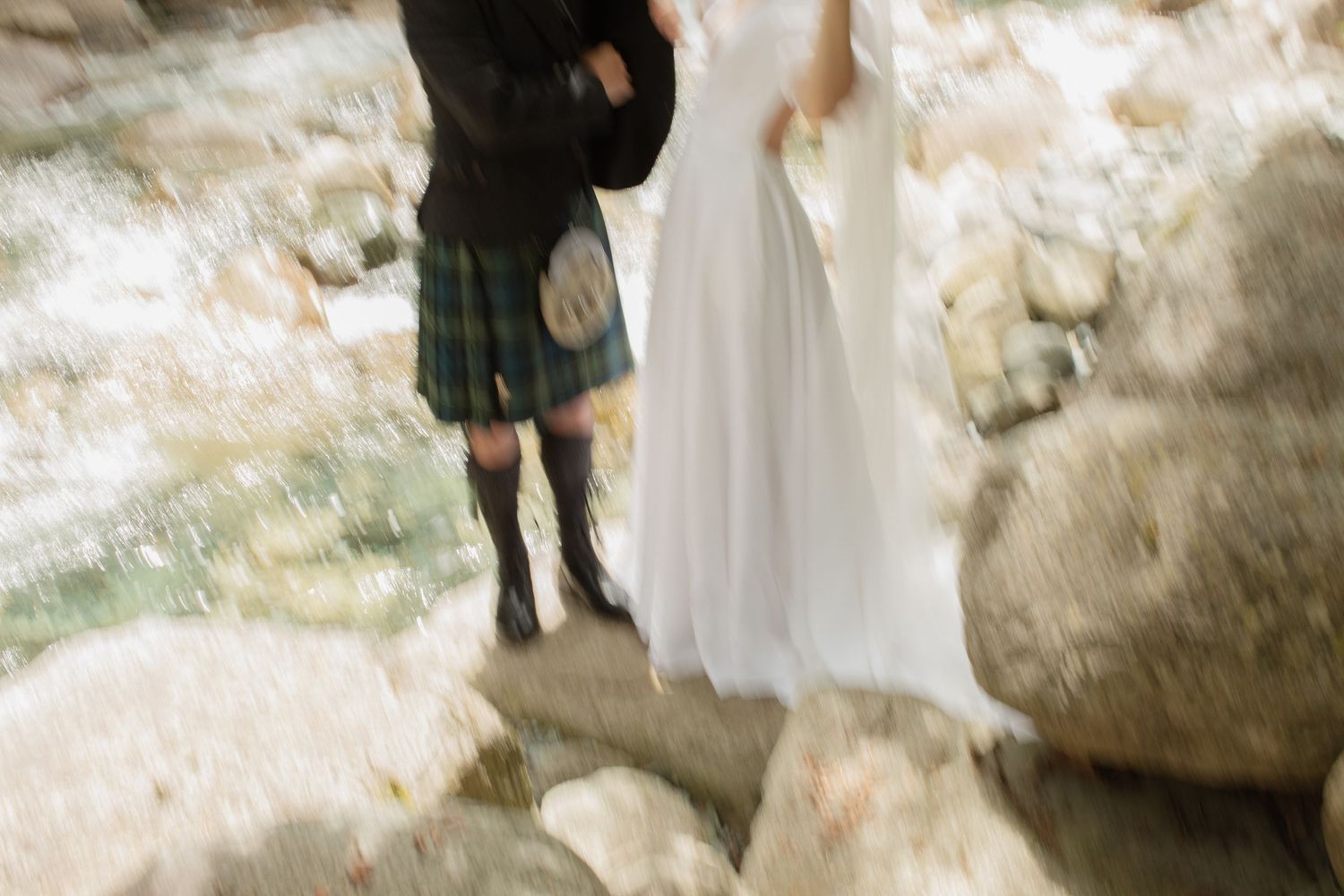 Groom in traditional Scottish tartan kilt standing with bride on rocky riverbank in forest setting.
