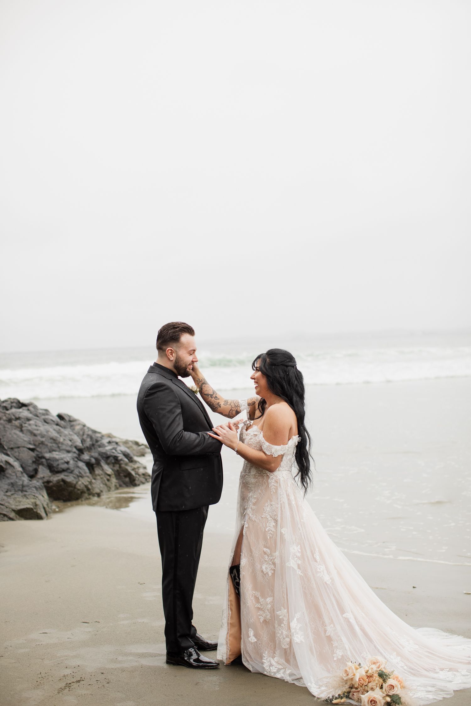 Bride in lace gown and groom in black suit hold hands facing each other on foggy beach.