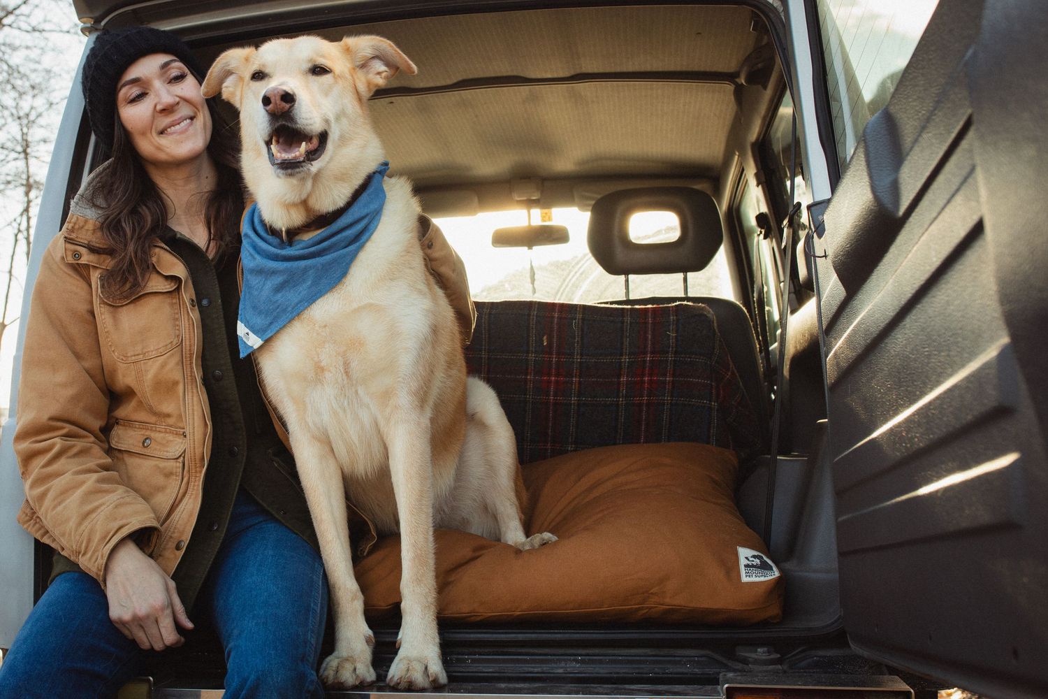 Smiling woman sitting in open car trunk with a happy yellow Labrador wearing a blue bandana.