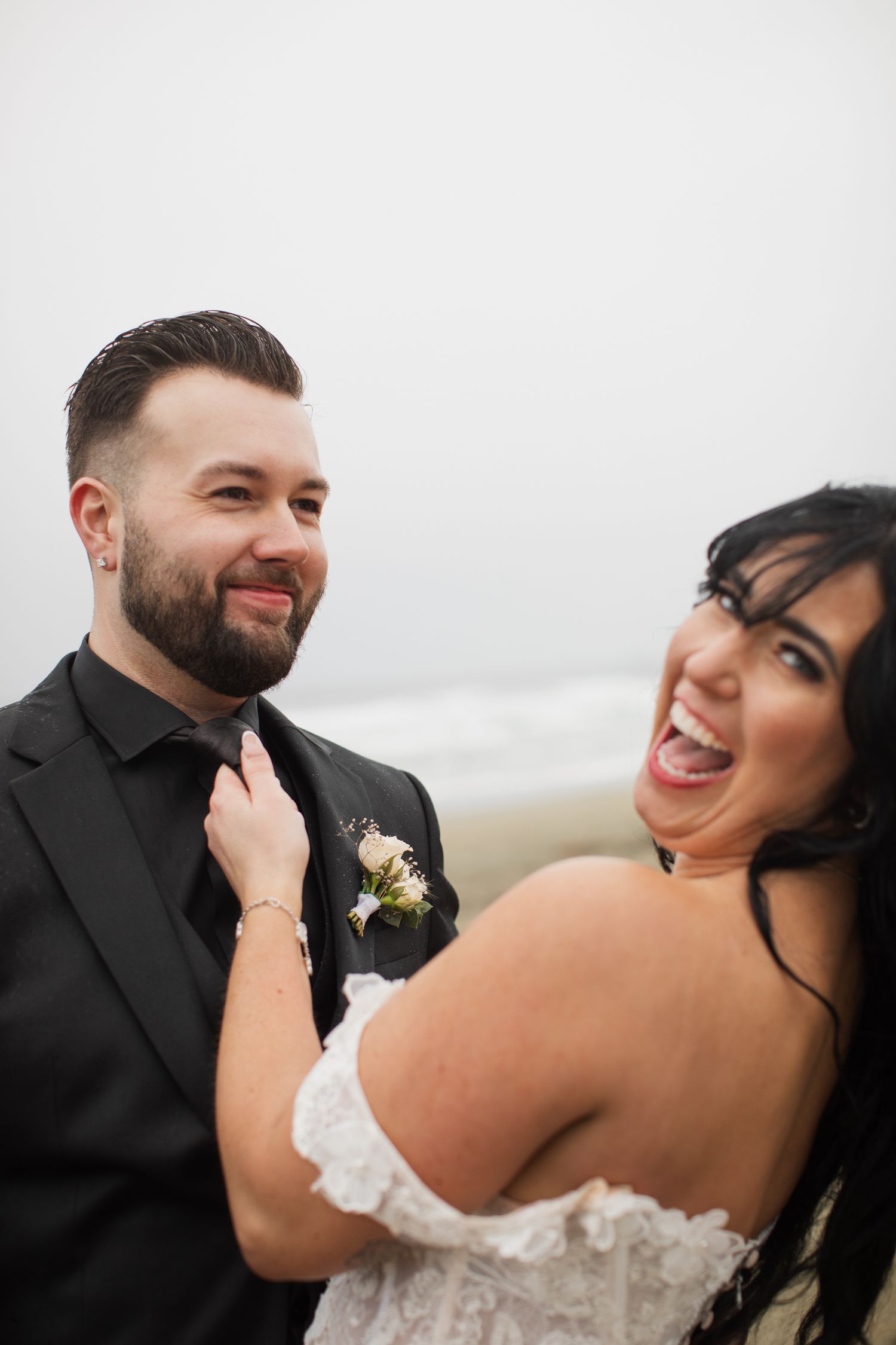 Laughing bride adjusts grooms black tie on foggy beach, both sharing a joyful candid moment.